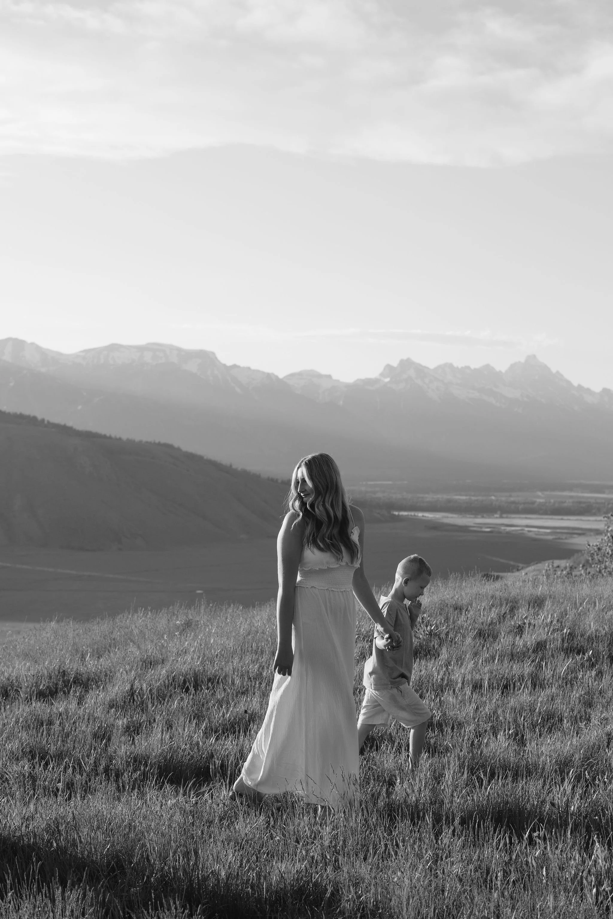 Sister holds younger brother's hand as they walk through grassy field during the Grand Teton sunrise.