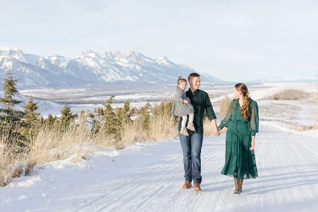 Mother and father walk hand in hand on a snow covered trail as they hold their daughter during a holiday card photoshoot.