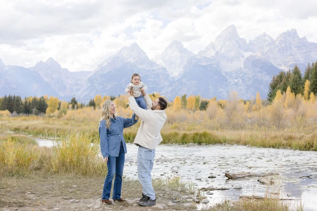 Father lifting baby in the air with mother smiling nearby in Grand Teton National Park with golden fall trees and mountain views