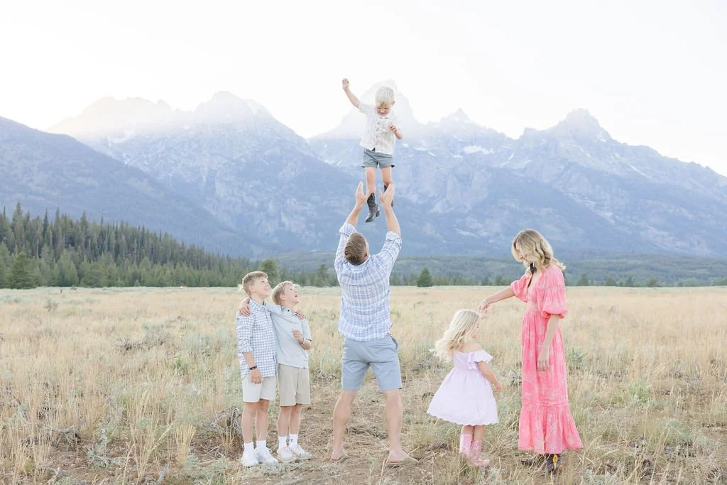 Father lifting child in the air while family plays in a mountain meadow with the Tetons behind them at Jenny Lake Grand Teton