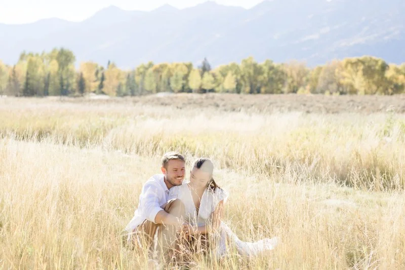 Couple sitting together in a golden field after their Jackson Hole proposal surrounded by breathtaking mountain views and romantic places to propose in the Tetons