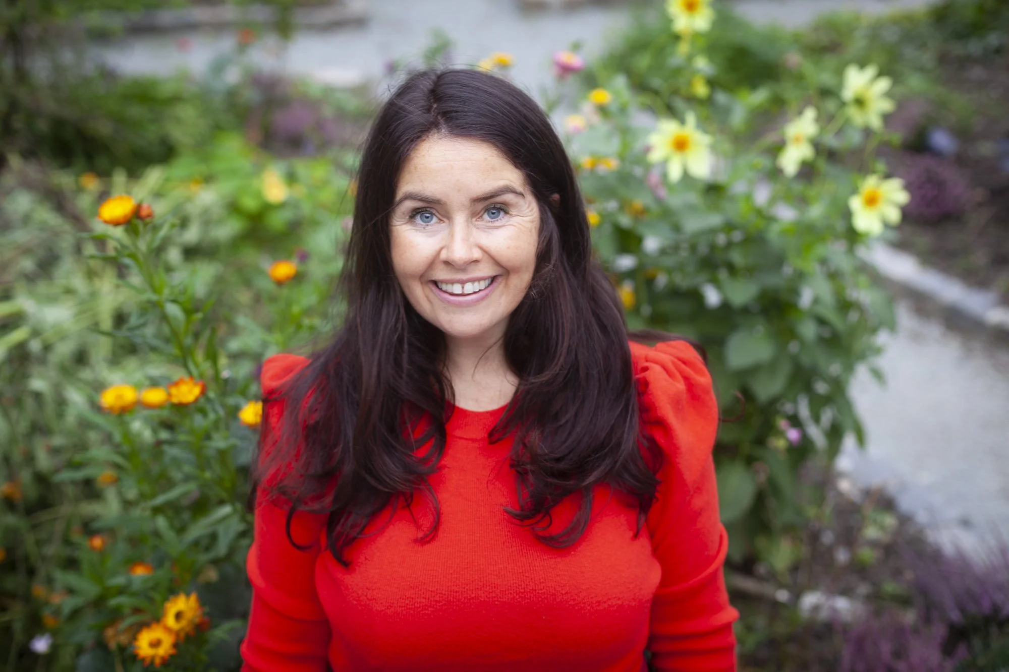 En kvinne med brunt hår og blå øyne smiler foran blomster i en hage, iført en rød genser.