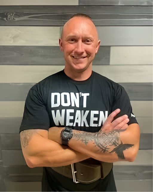 Man with tattooed arms crossed over his chest, wearing a red Tshirt and smiling.