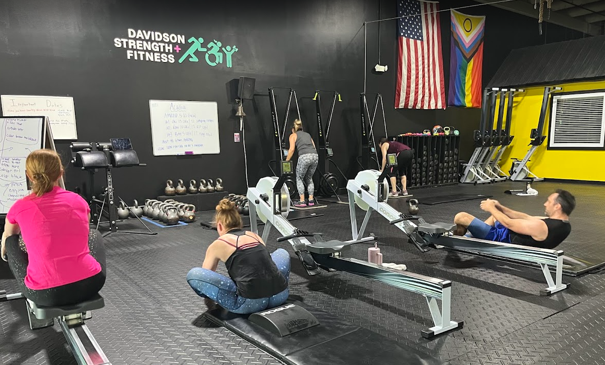 3 people on rowing machines in a gym with an American flag and a PRIDE flag on the wall.