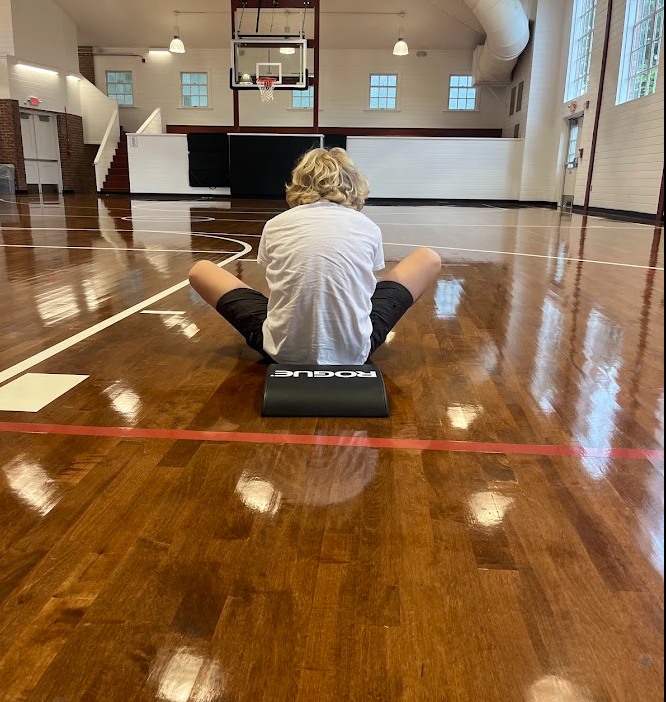 Picture of the back a school age boy in a white Tshirt, seated, doing Sit-ups, in a large gymnasium with wood polished floor.