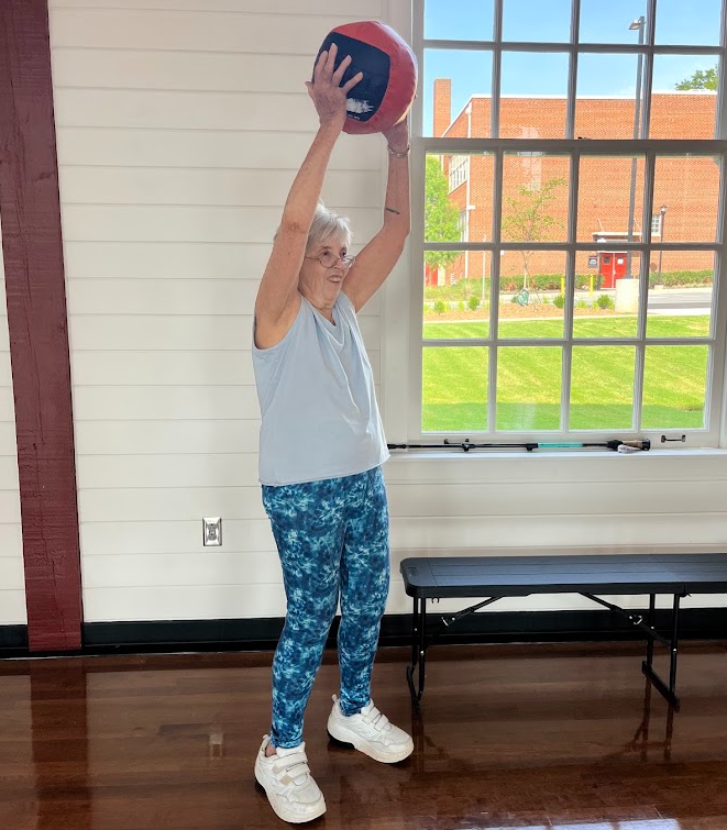 older woman lifting a ball over her head and smiling. performing deadlift