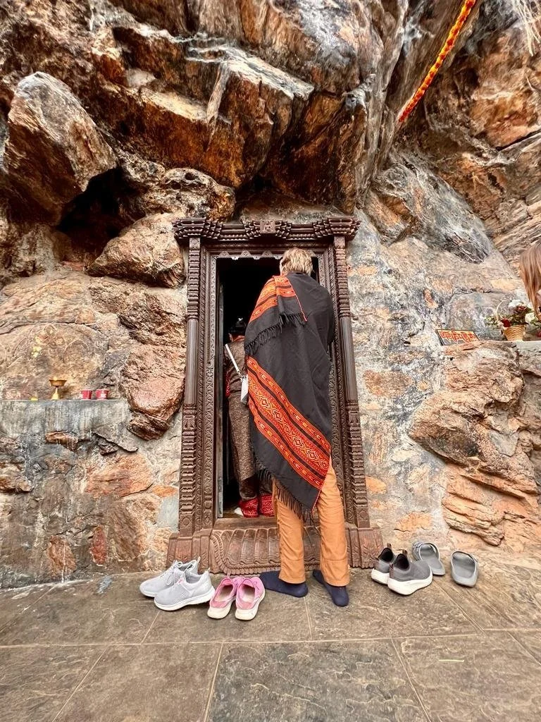 Two women in traditional Indian sarees standing and talking to a man sitting on a bench in a park with ruins and a white domed building in the background.