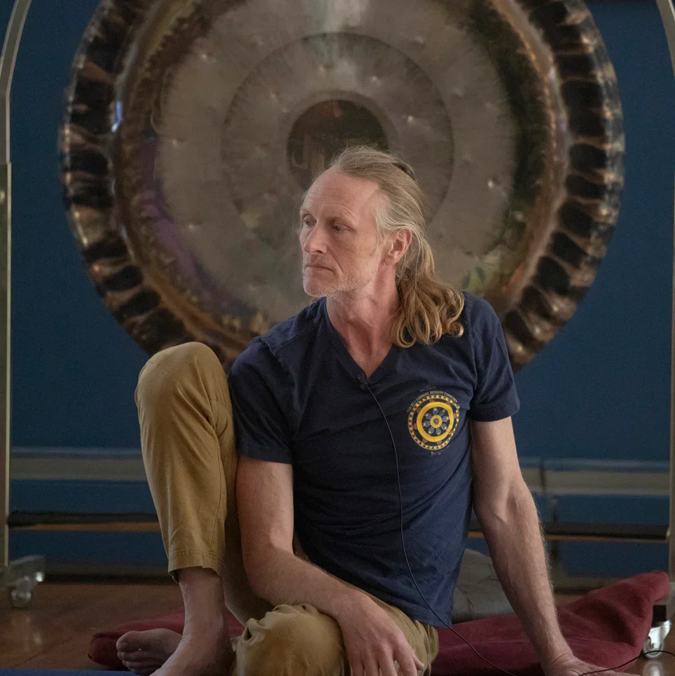People practicing yoga in a studio, with a male instructor assisting a woman in a yoga pose, others lying on mats in the background.