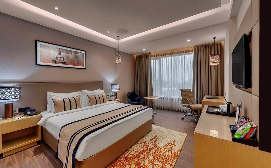 Hotel room with a bed, wooden wall accents, a small television, air conditioning, window with brown curtains, and ceiling with recessed lighting and a fan.
