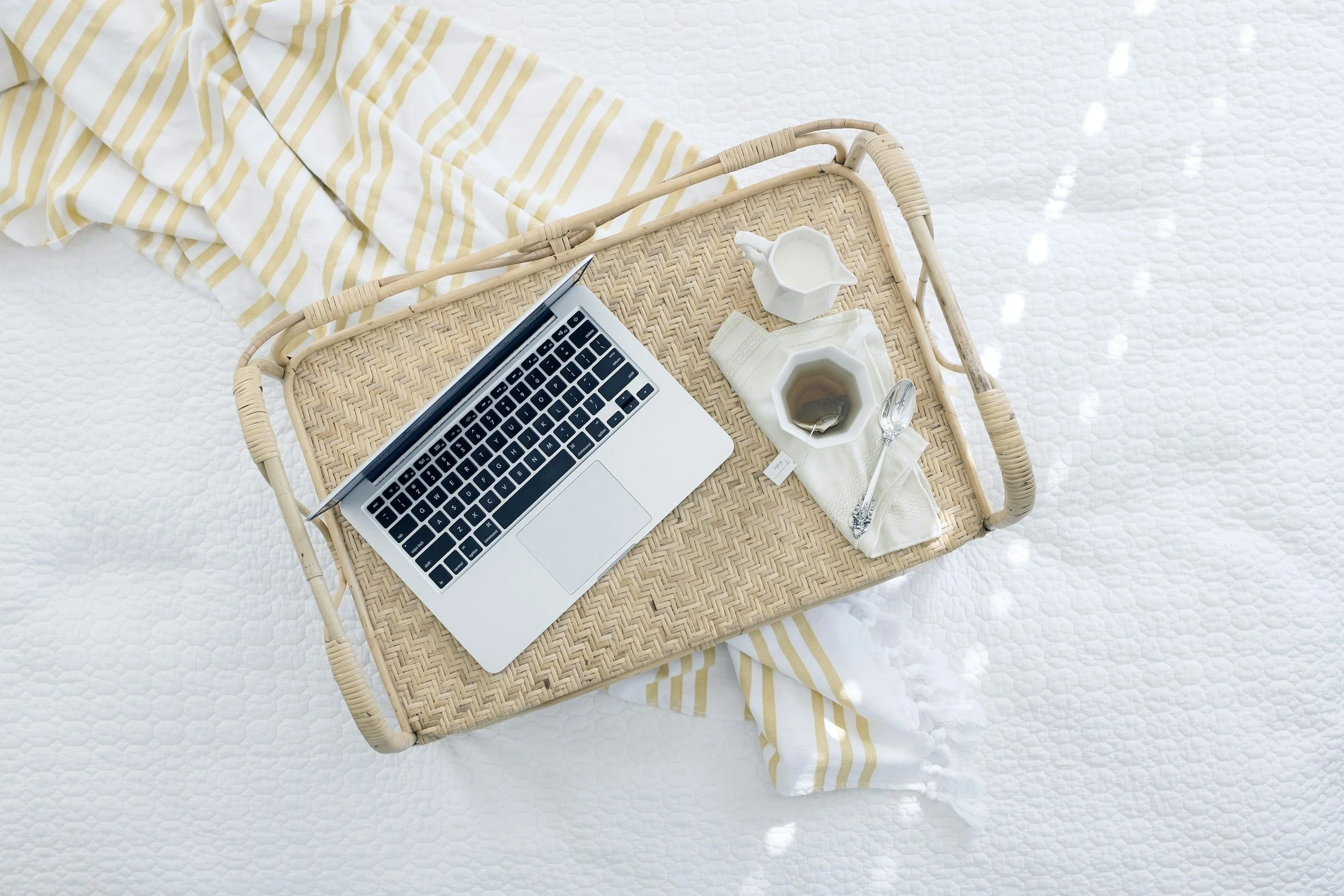 Wicker tray with a laptop, a white creamer, a cup of tea, a spoon, and a napkin on a bed with white bedding and a yellow striped blanket.