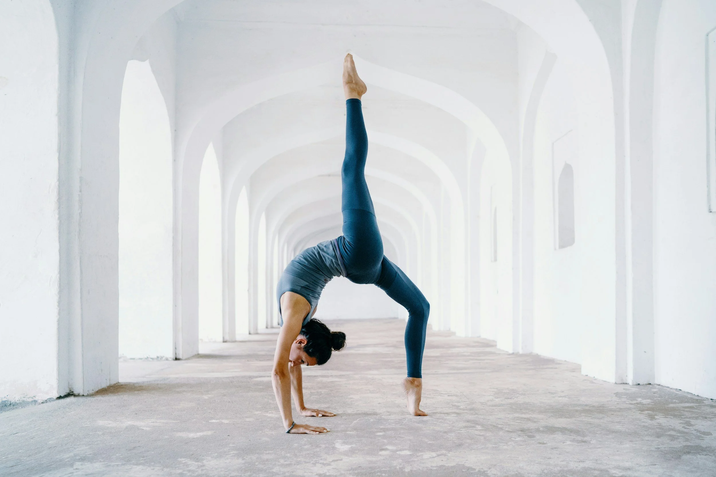 A woman practicing yoga in a handstand split pose in a bright, white hallway with arches.
