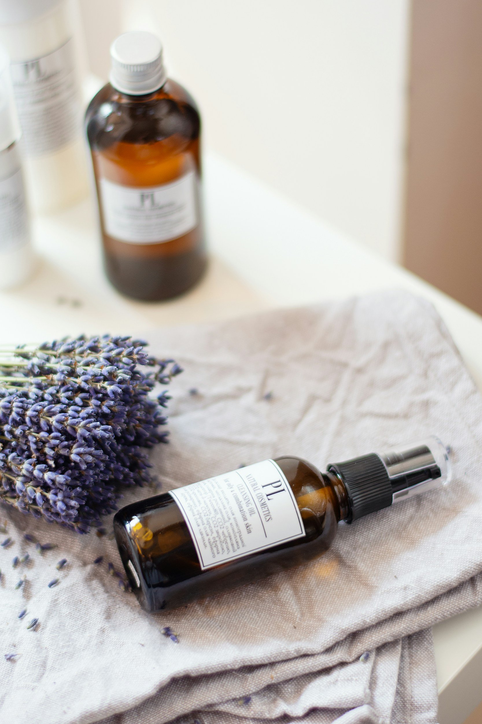 A spray bottle of lavender oil next to a bunch of fresh lavender and several other bottles on a white surface.
