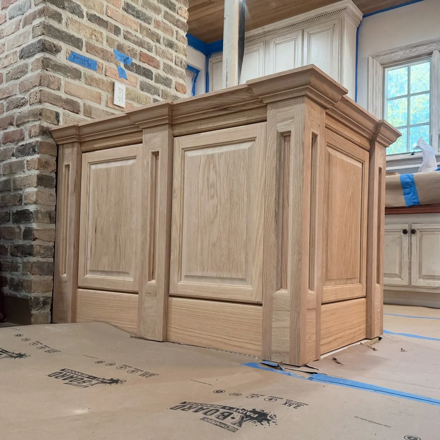 Unfinished wooden kitchen cabinet or island with decorative molding in a kitchen under construction, with a brick wall, window, and protective floor covering visible.