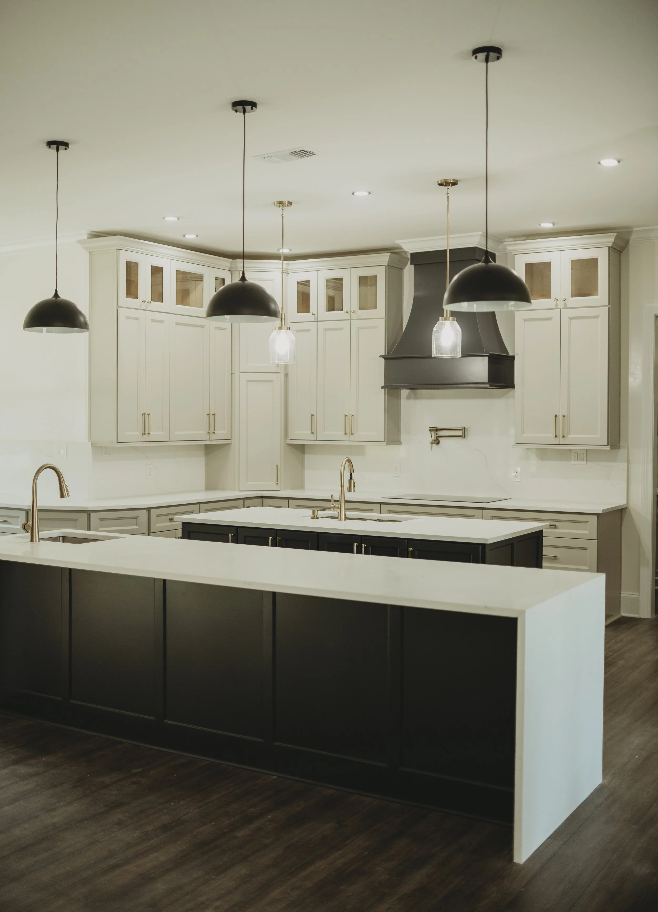 Modern kitchen with a white island, black cabinetry, white cabinets with gold handles, pendant lights, and a range hood, with dark hardwood flooring.