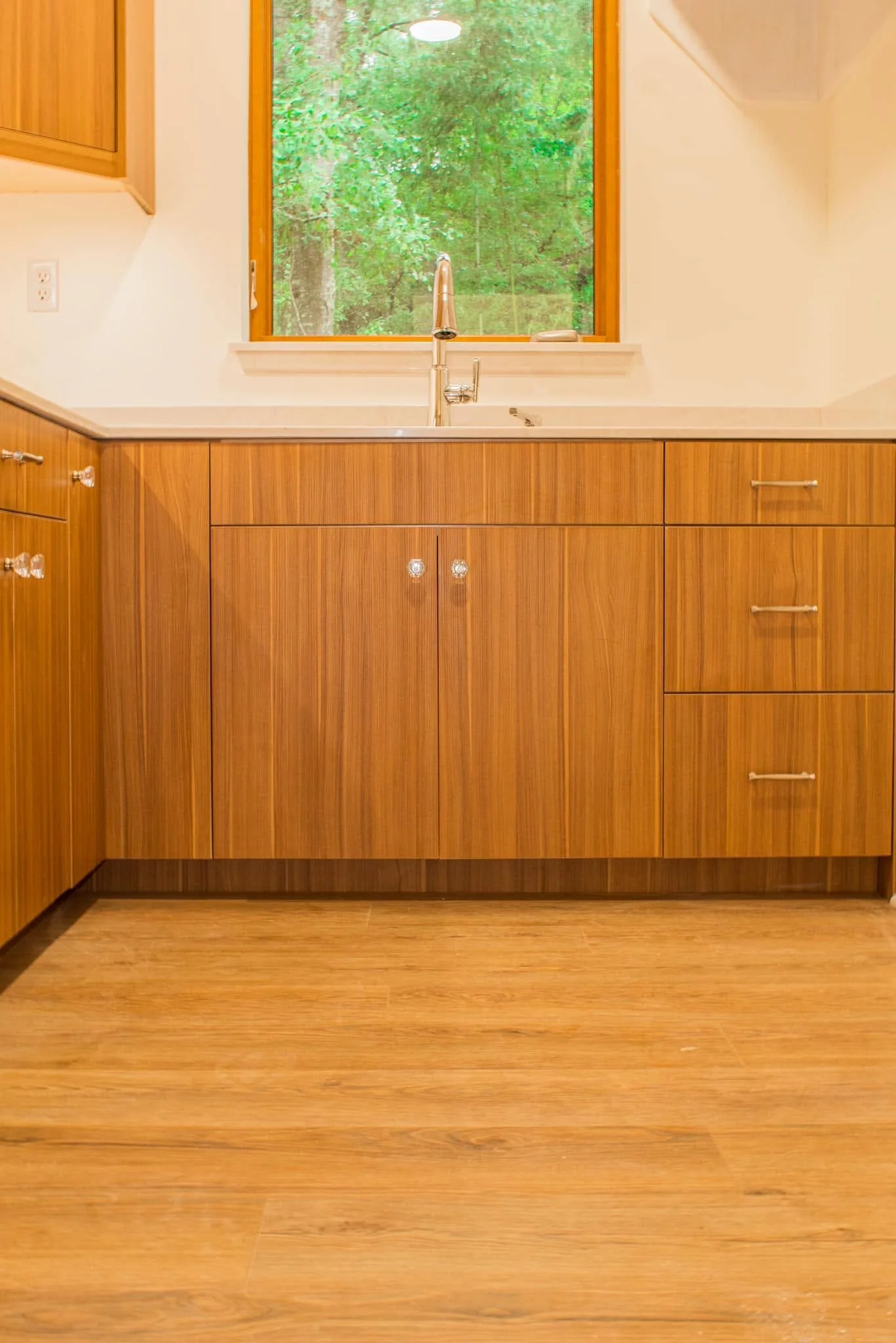 View of a kitchen with wooden cabinets and flooring, a window overlooking green trees, and a modern silver faucet above a white countertop.