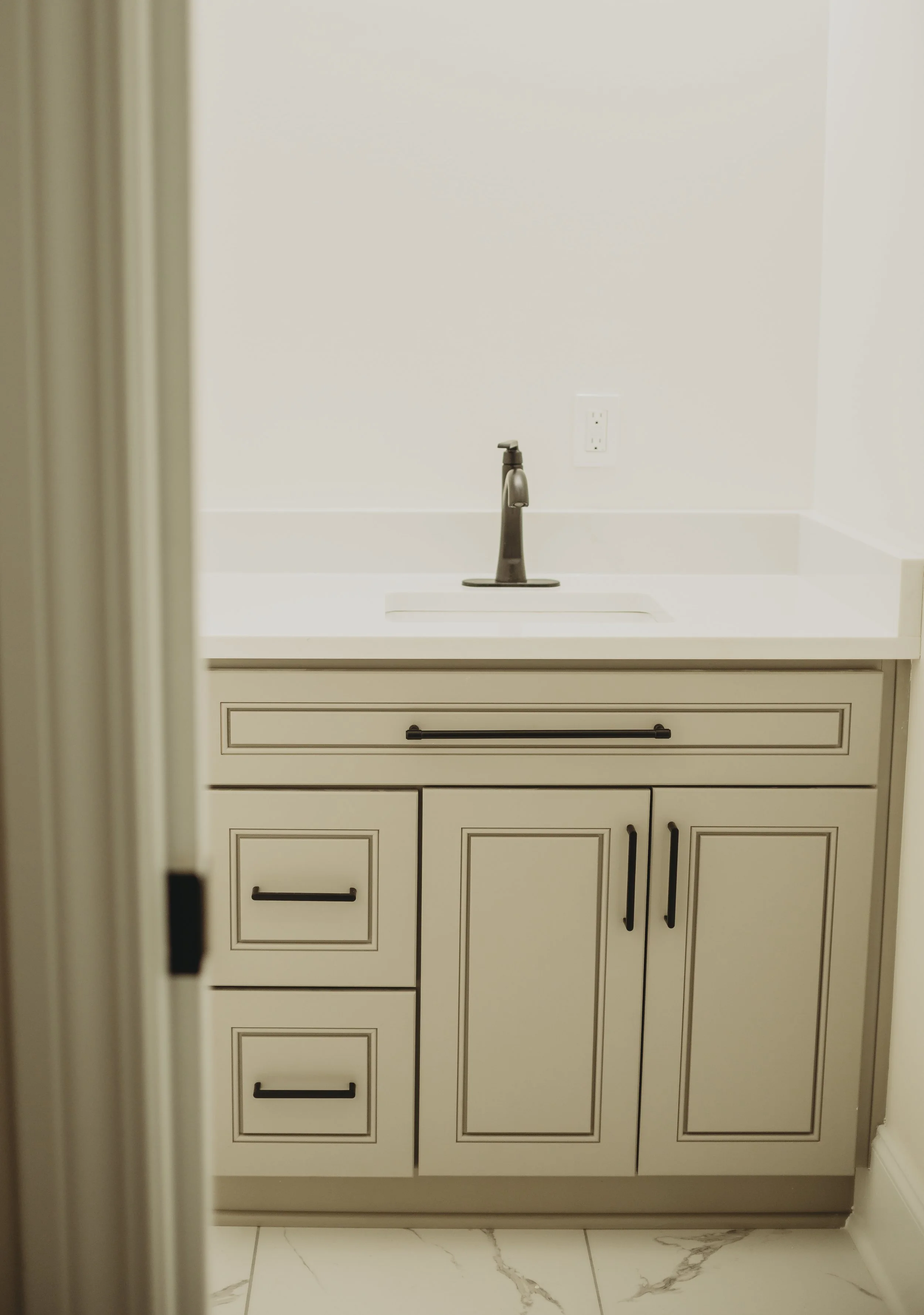 Bathroom vanity with a silver faucet and beige cabinet with black handles, set against a plain white wall, with a power outlet visible behind the faucet.