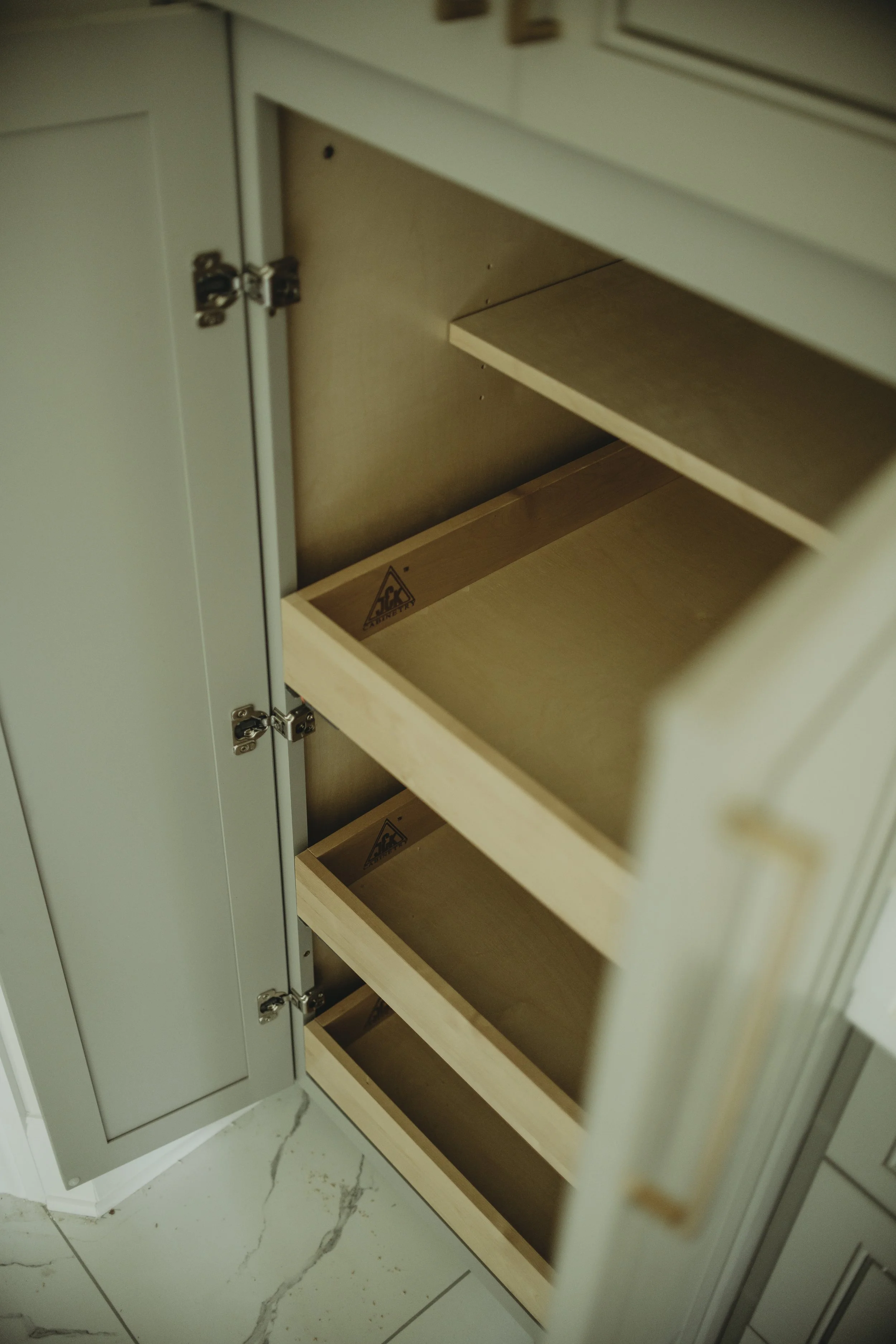 An empty kitchen cabinet with multiple shelves, partially open door, and metal hinges. The cabinet appears to be made of light wood or laminate, with a sticker or label at the bottom shelf. The floor is tiled with white marble pattern.