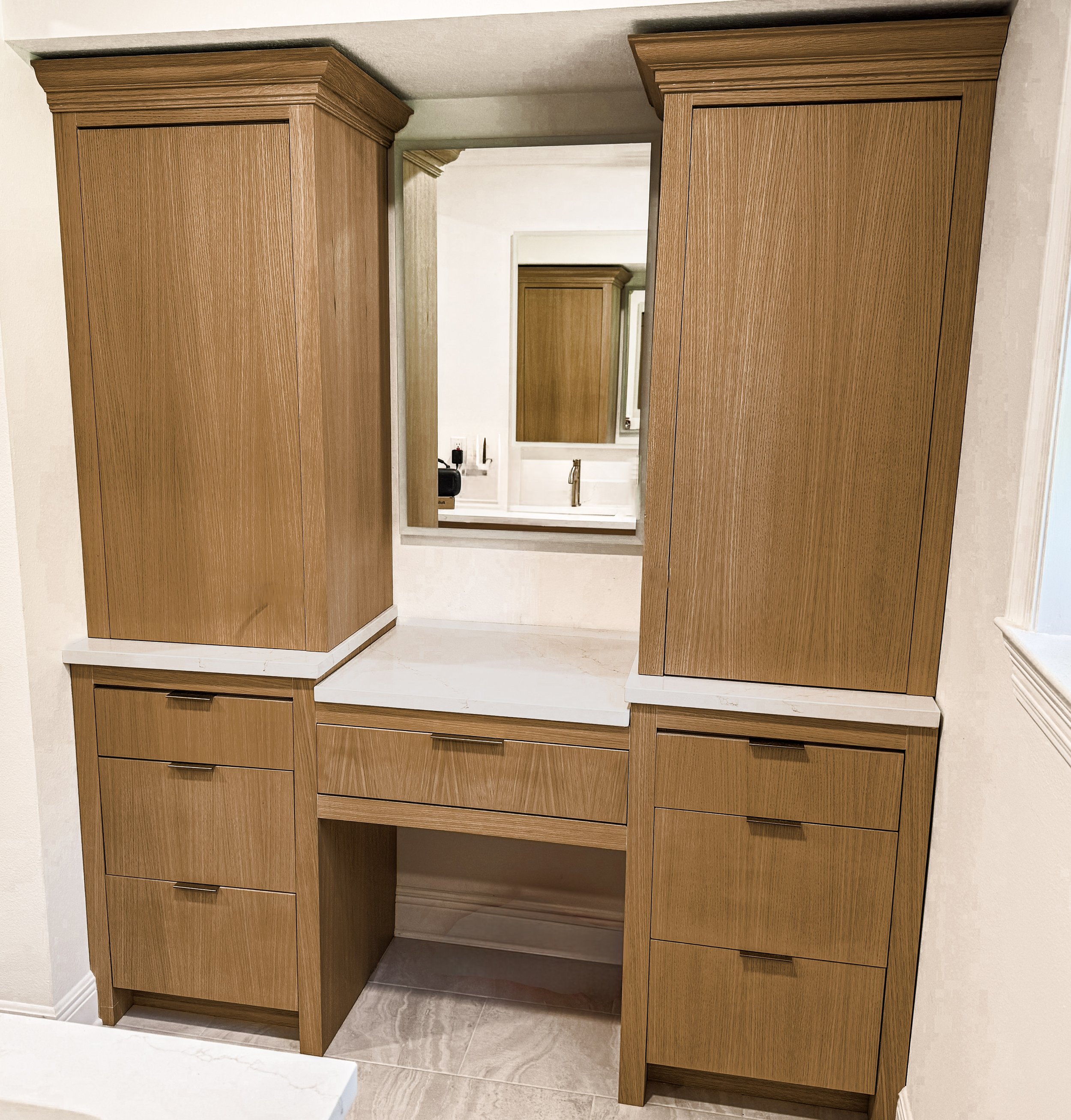 Wooden bathroom vanity with multiple drawers and cabinets, mirror behind, and a view of a sink and wall-mounted mirror in the reflection.