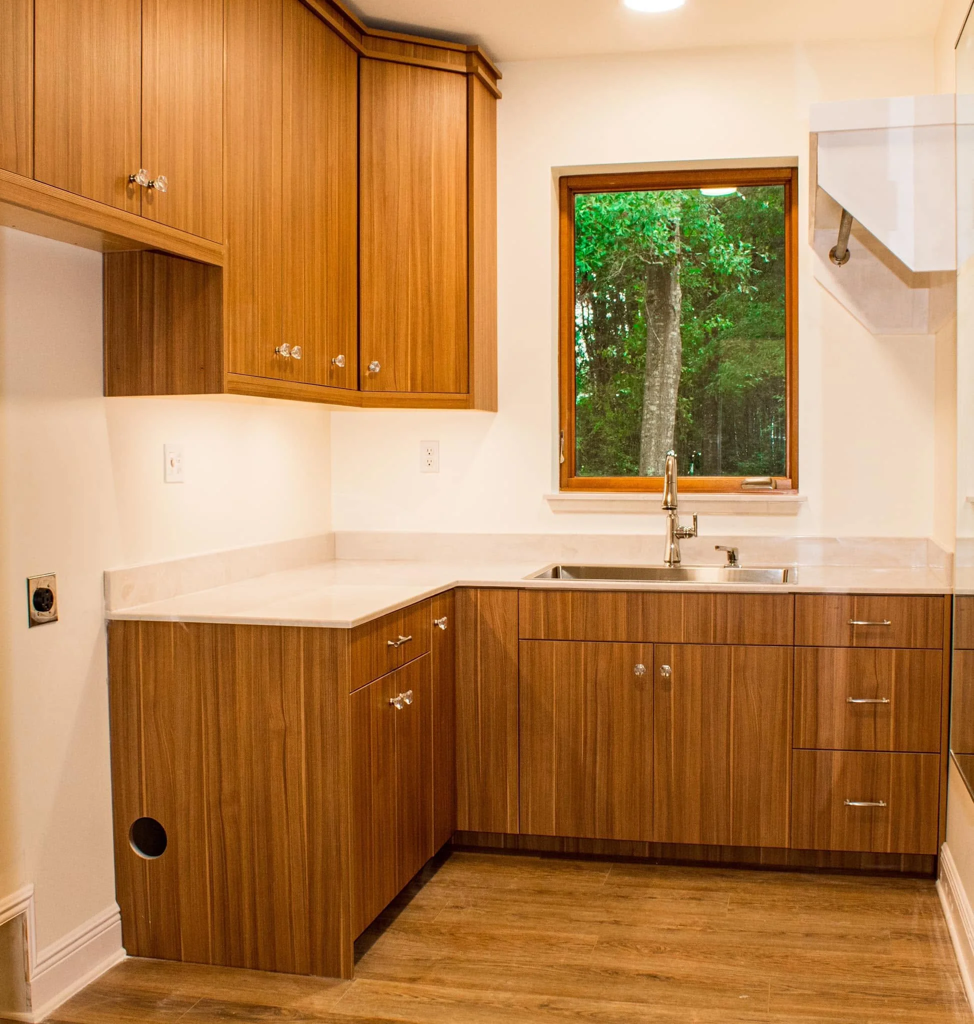 Kitchen with wooden cabinets, a window showing trees outside, a sink under the window, and a white countertop.