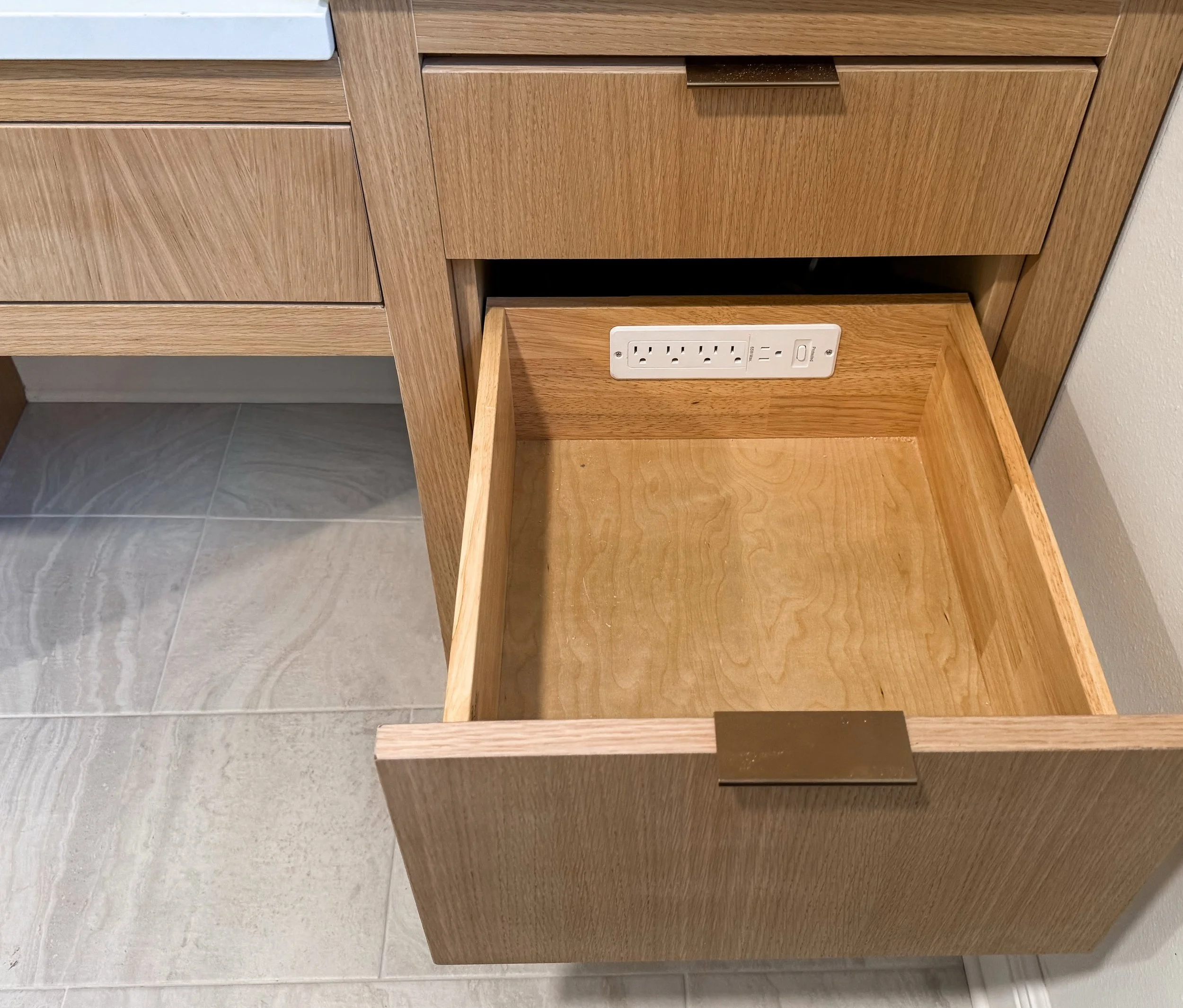 A wooden nightstand with two drawers, one partially open showing an electrical outlet panel inside the drawer. The nightstand is next to a section of wooden cabinetry and a white wall. The outlets include two three-prong sockets and switches.