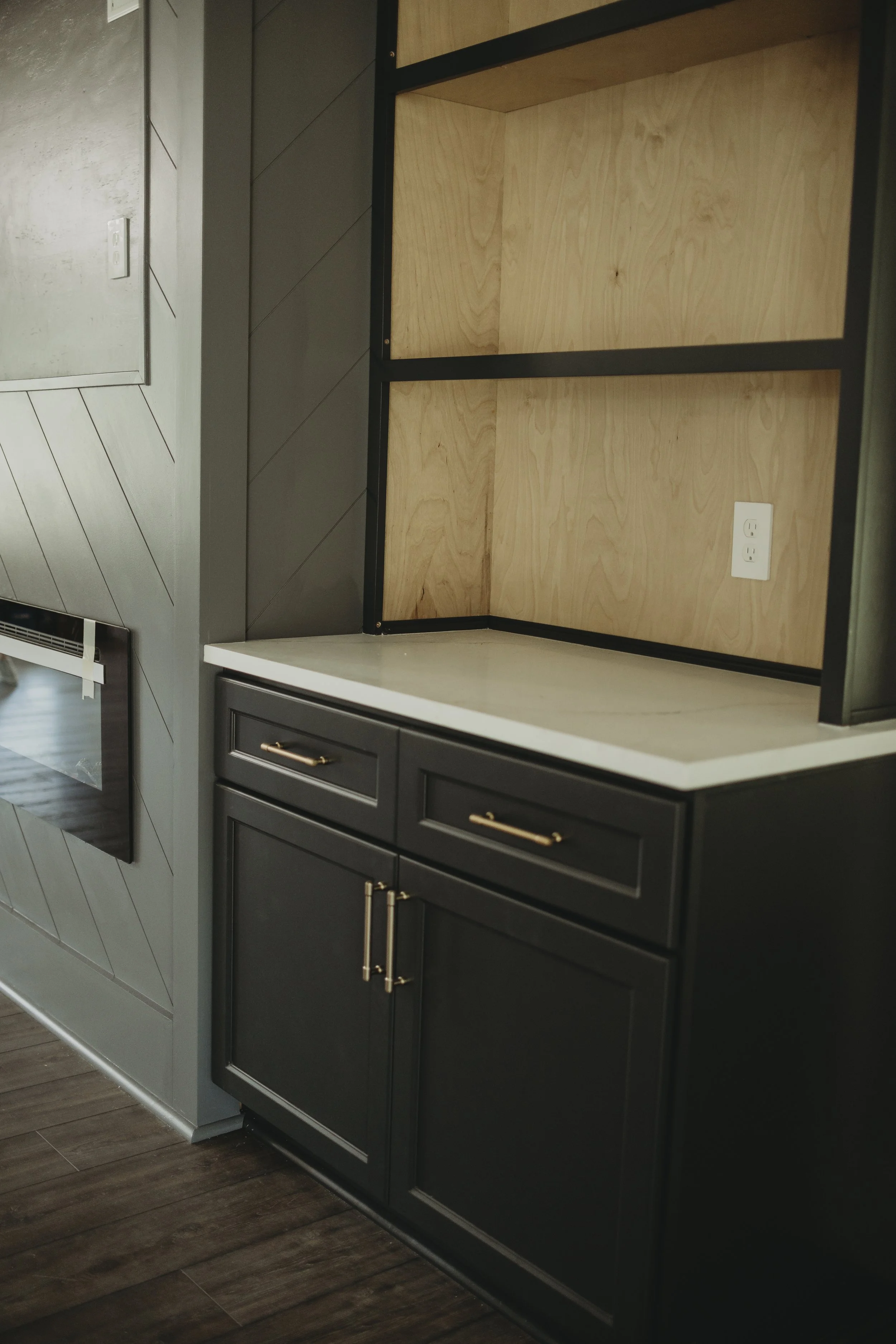 Empty kitchen cabinet with black frame and wood interior, white electrical outlet on wood wall, adjacent dark gray cabinetry, and hardwood floor.