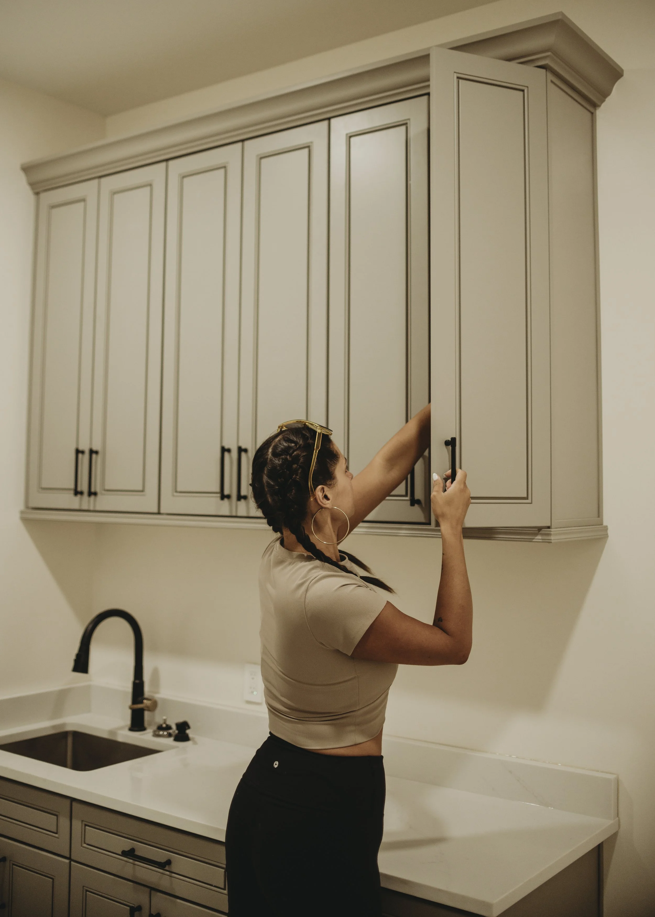 A woman with braided hair and hoop earrings is taking a photo with her phone in a kitchen. The kitchen has a marble countertop, a black faucet, beige cabinets, and a beige wall.