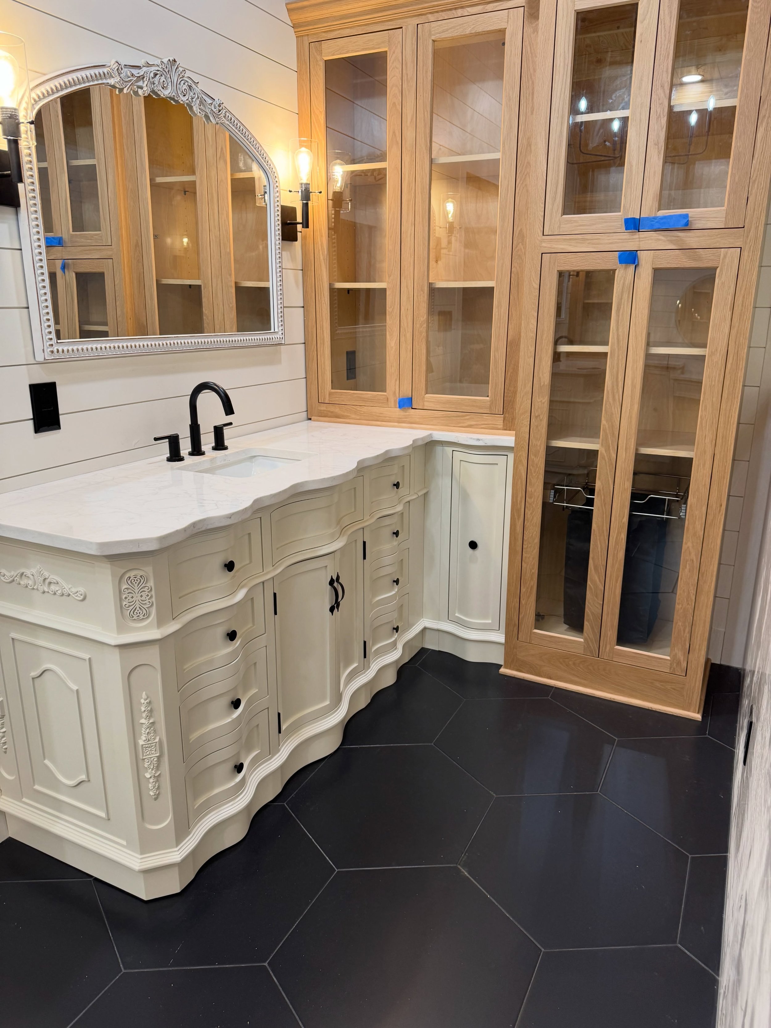 A bathroom vanity with a white ornate cabinet, a black faucet, and a marble countertop. A large mirror with a decorative frame hangs above the sink. To the right, there is a tall wooden cabinet with glass doors, blue painter's tape on the edges, and 