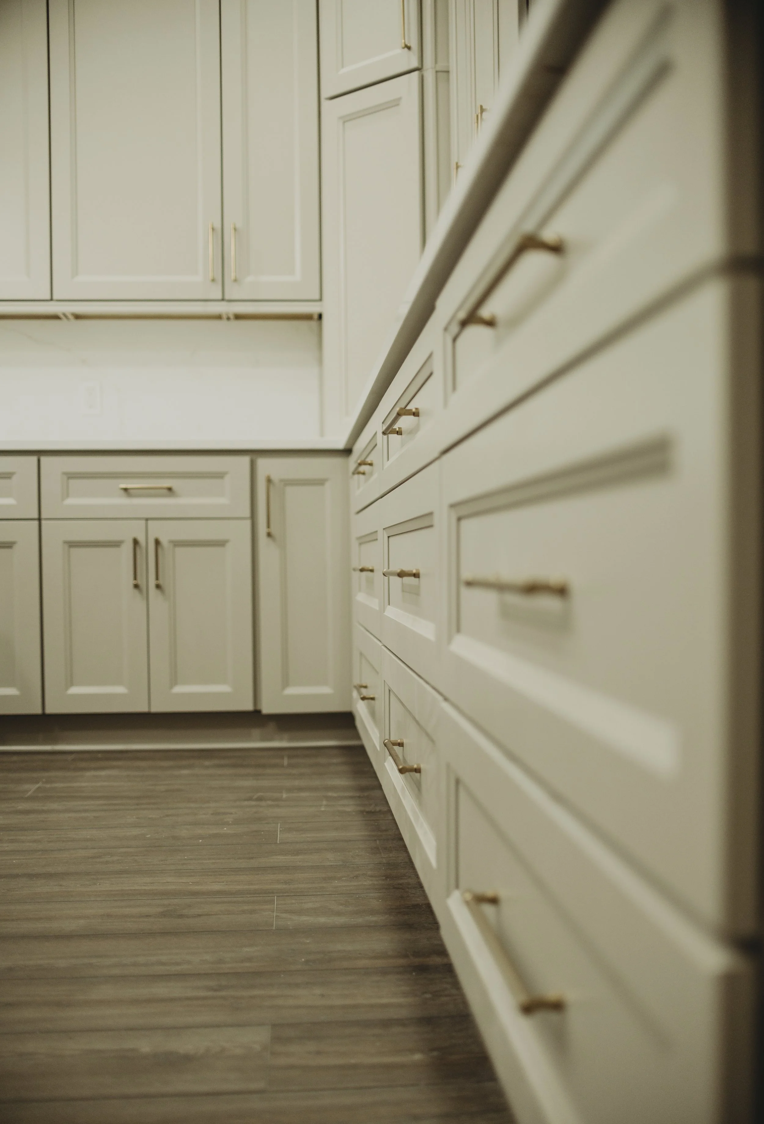 A kitchen with cream-colored cabinets and drawers, gold handles, and wood-look flooring. The cabinets are arranged along two walls, with some cabinets above and some below the countertops.
