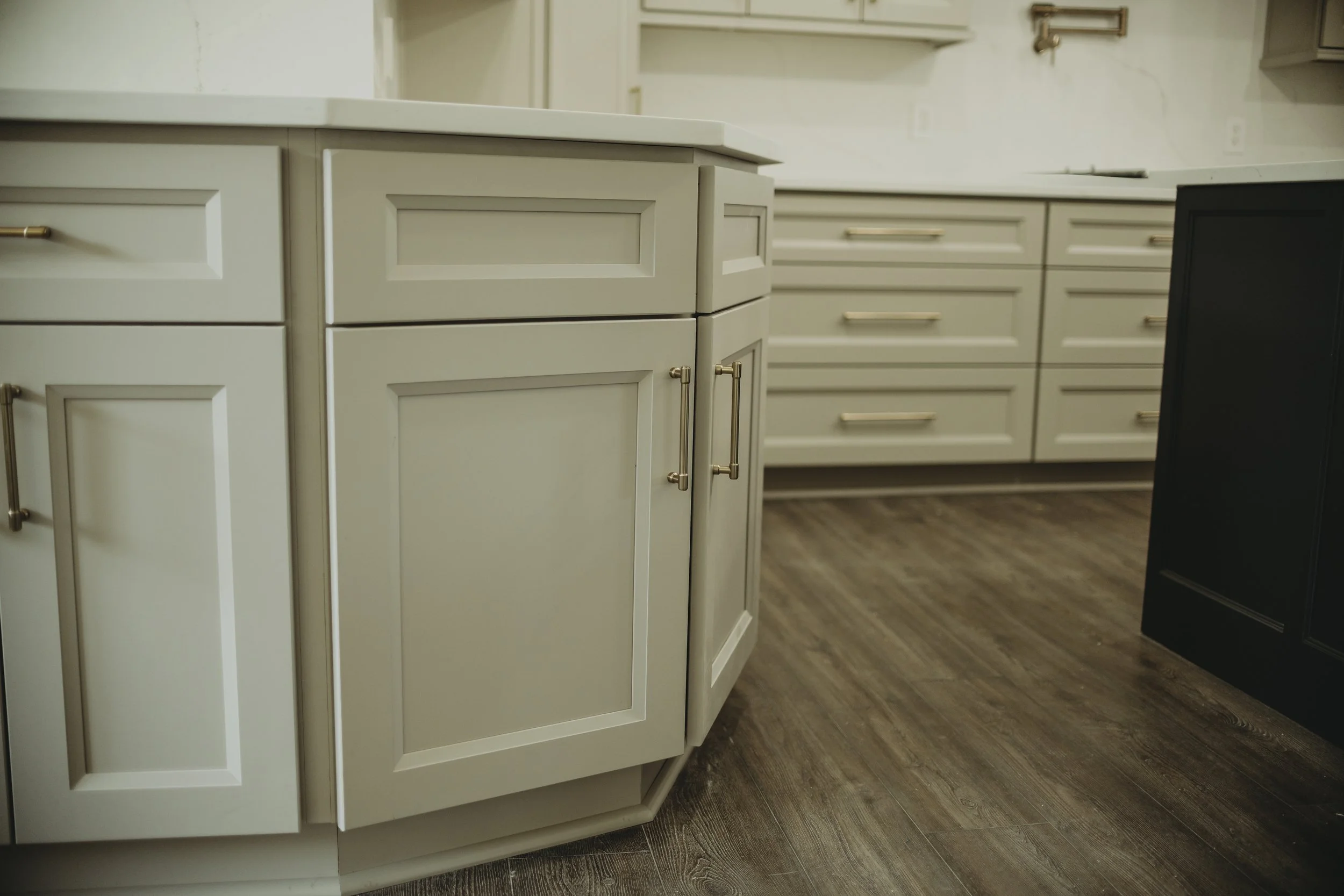 Close-up of light-colored kitchen cabinets with gold handles and a darker cabinet on the right, on a wood-like floor.