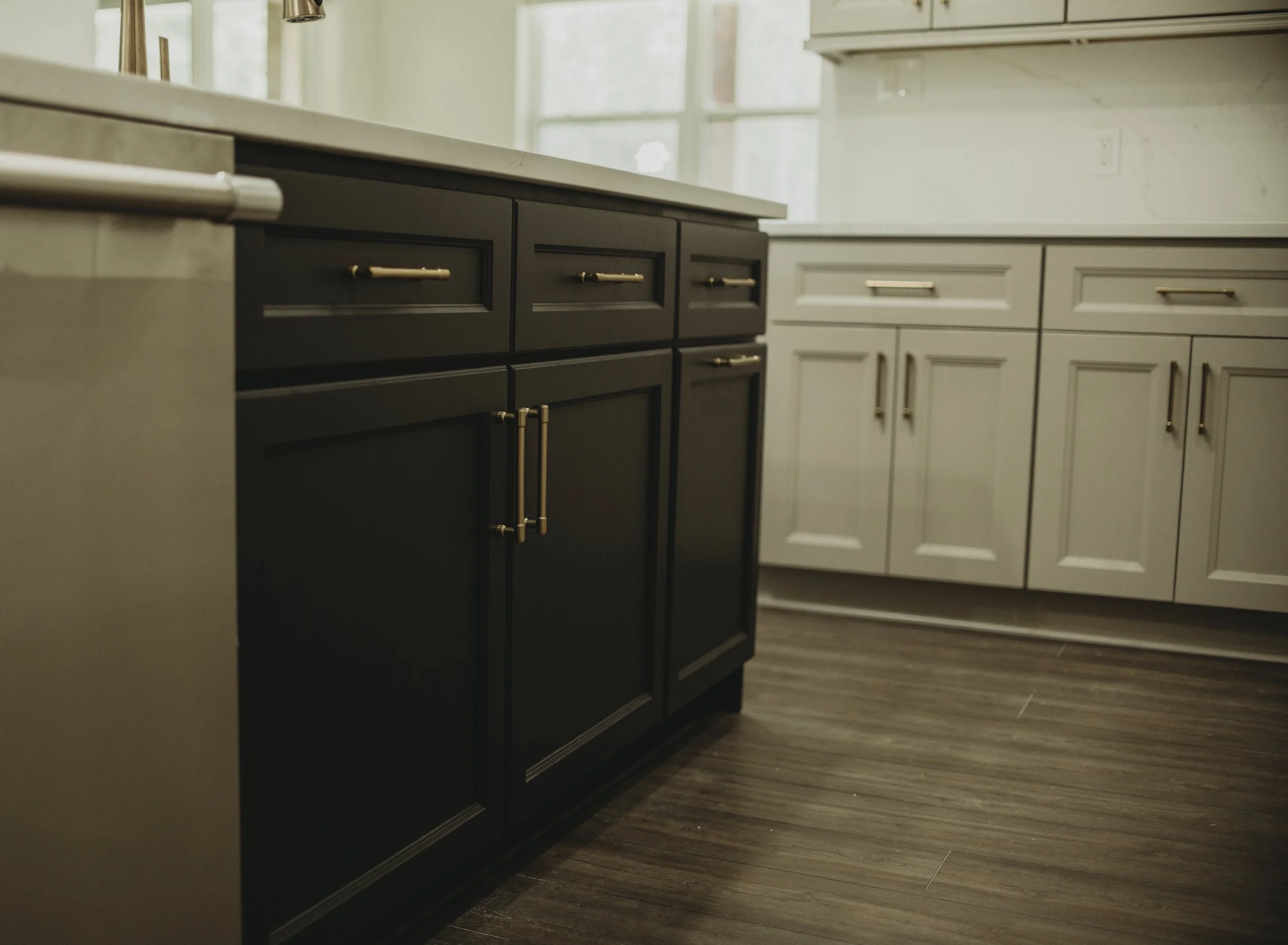 Kitchen with dark and light cabinetry, stainless steel handles, wooden flooring, and white walls with large windows.