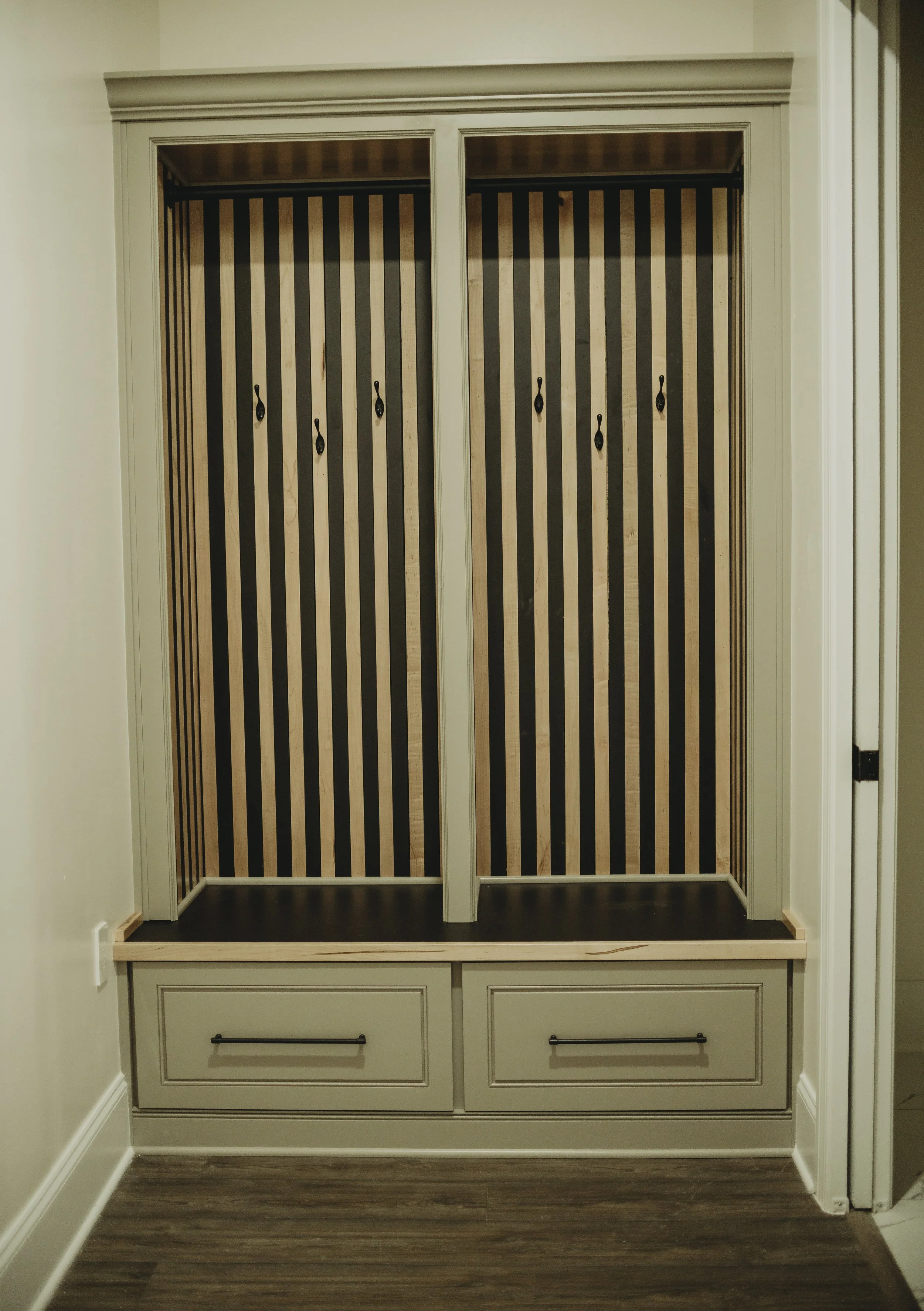 Custom built-in mudroom with hooks featuring vertical striped black and beige panels, and two drawers at the bottom with black handles, set against a beige wall with white molding.