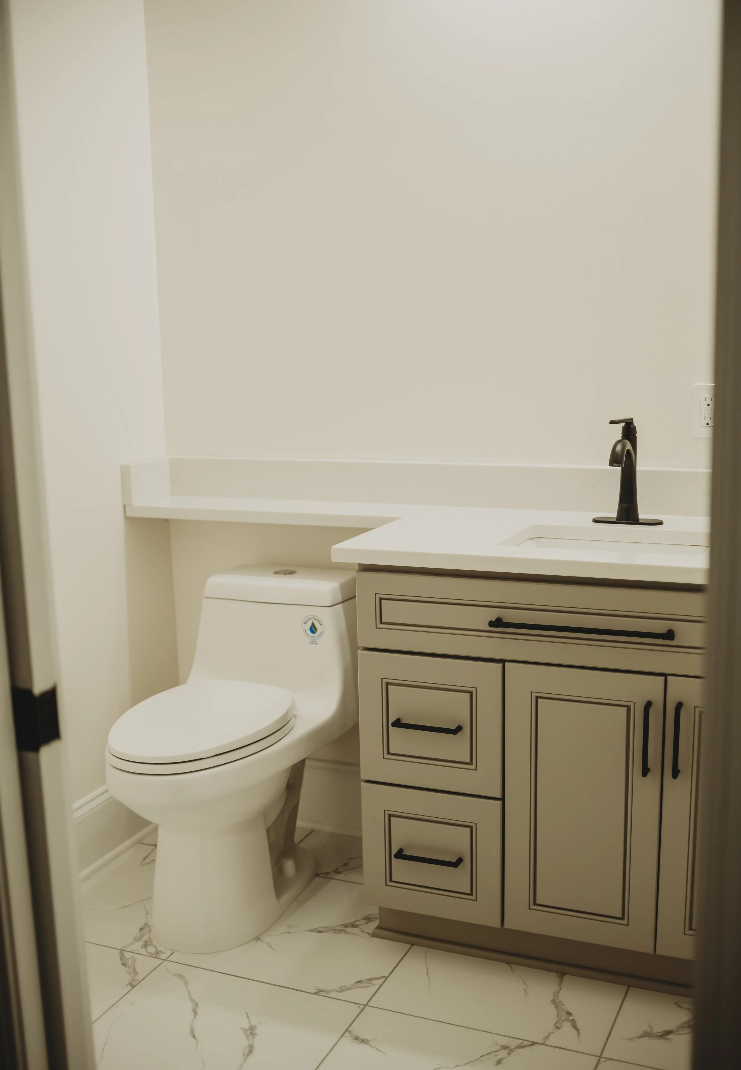 A bathroom with a white toilet, a beige cabinet with black handles, a white countertop with a black faucet, a wall outlet, and beige tiled flooring with dark veins.
