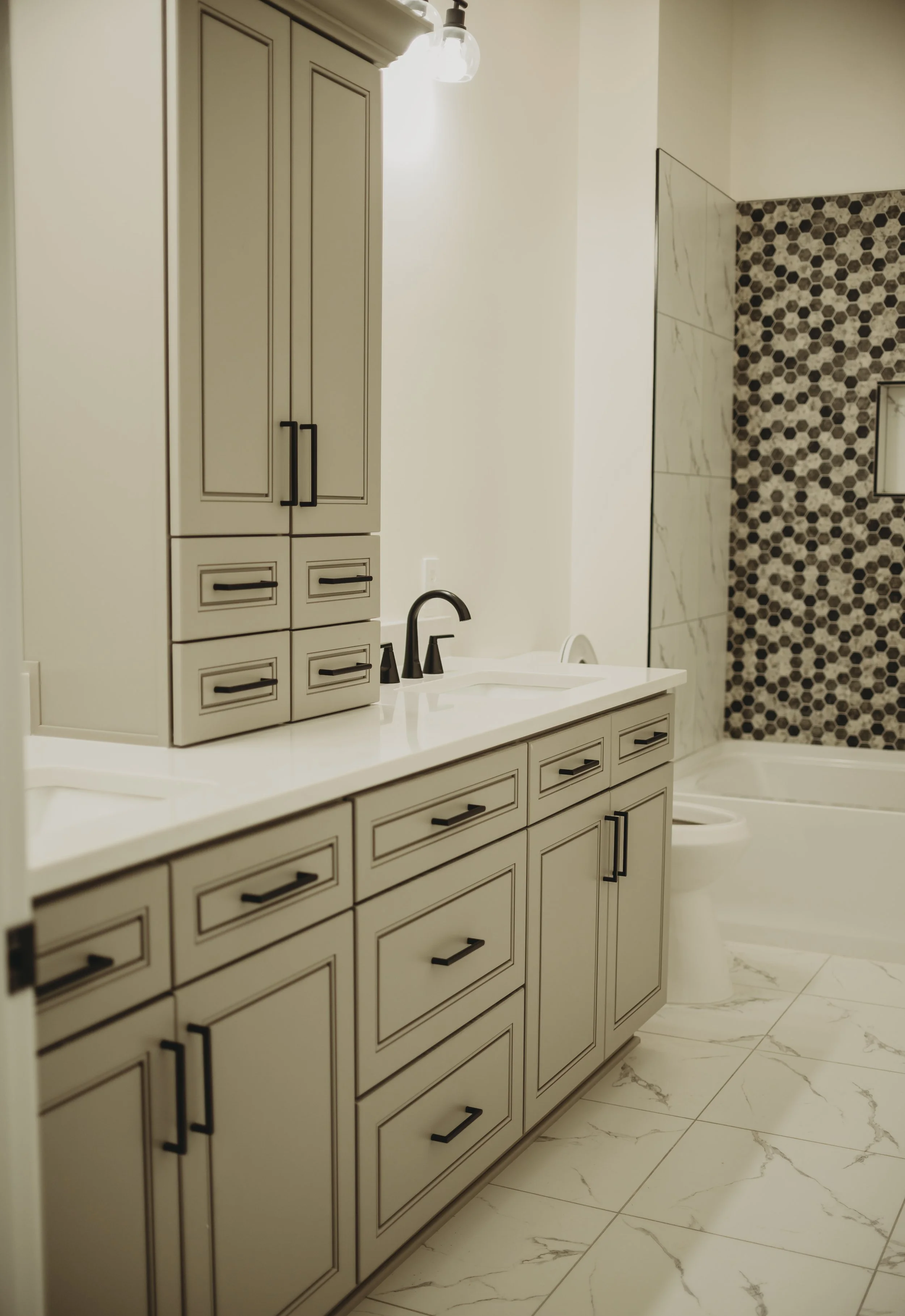 A bathroom with beige cabinets, black handles, a white marble countertop with a black faucet, a toilet, and a wall with a mix of white and honeycomb-patterned tiles.