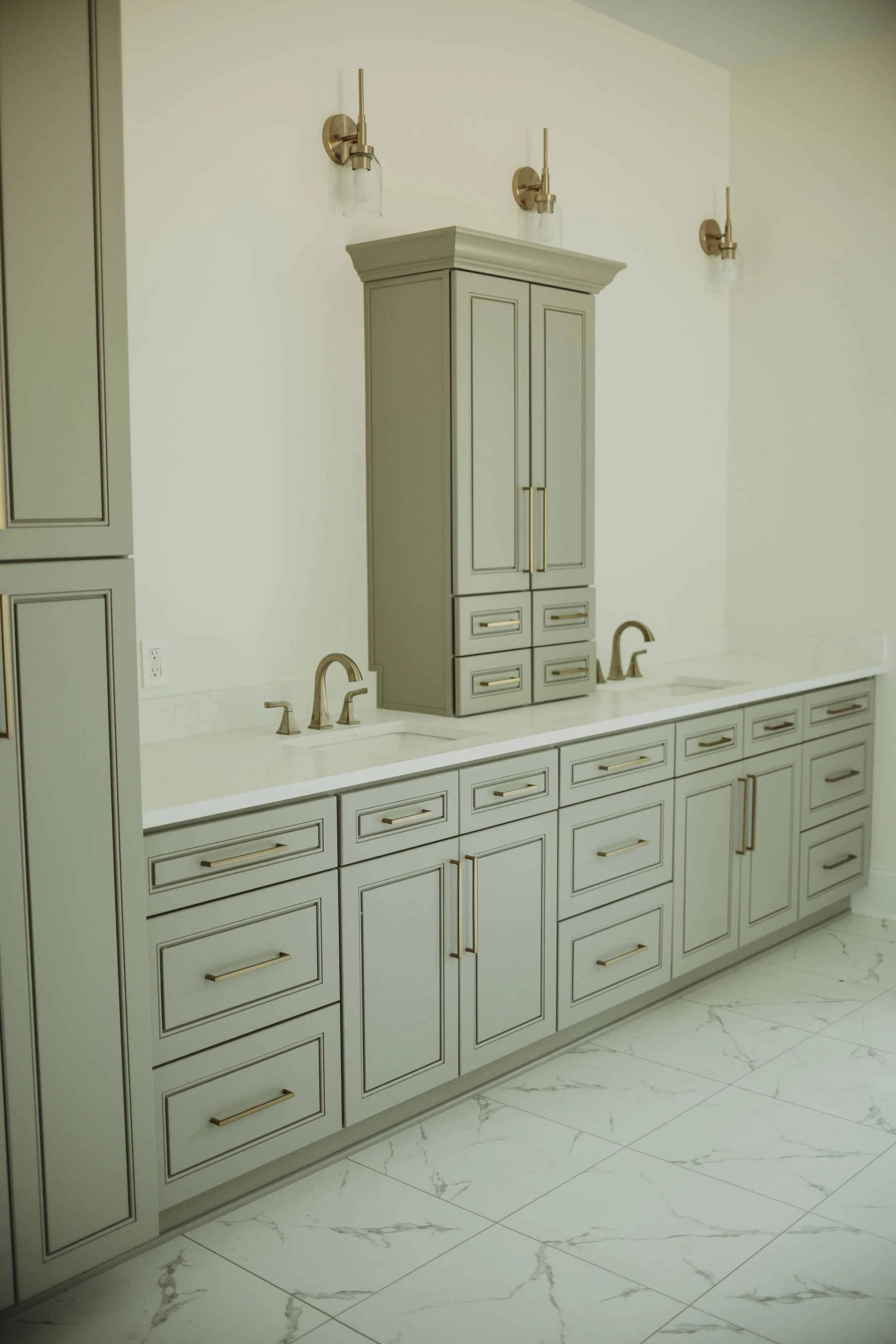 A bathroom or vanity area with light green cabinetry and gold hardware, featuring multiple drawers and cabinets below a white marble countertop. There are two sinks with gold faucets, and a tall matching cabinet in the center. The wall above has thre