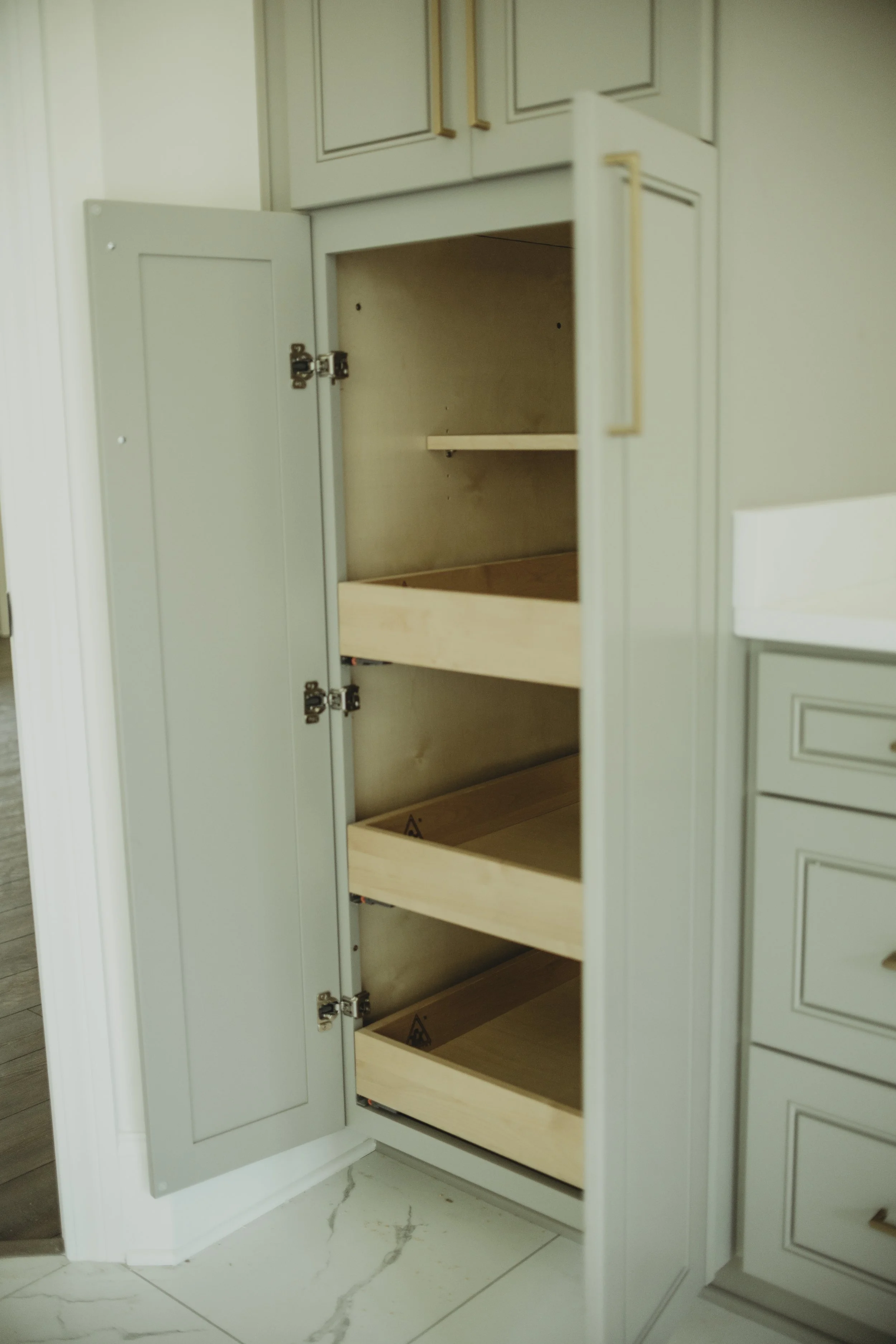 Open kitchen cabinet with light wood interior and empty shelves, adjacent to other cabinets, near a marble floor.