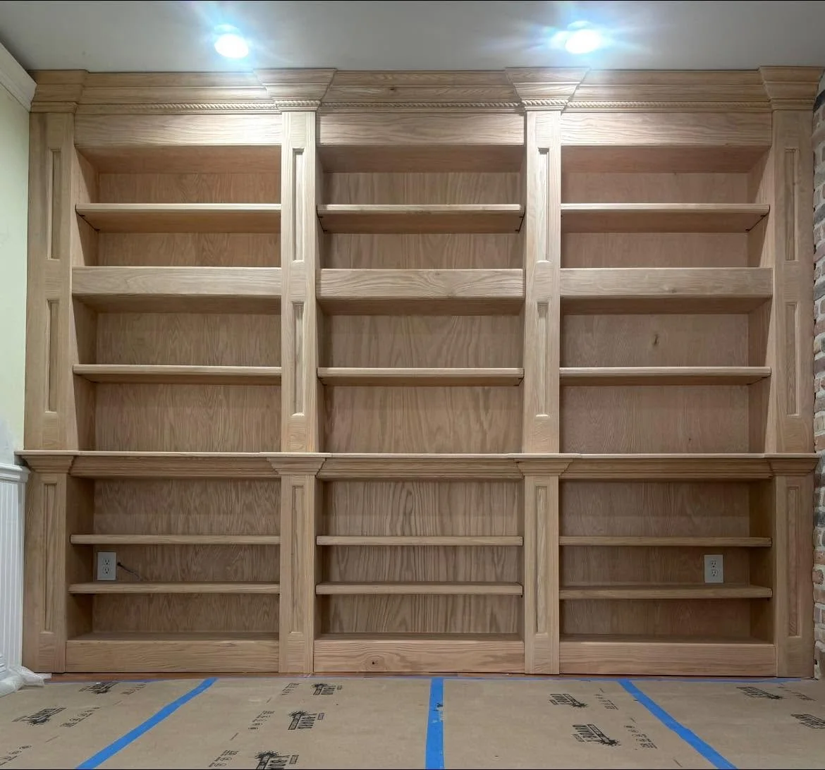 Empty wooden built-in bookshelf with three sections, multiple shelves, and electrical outlets at the bottom, against a wall with brick on one side.