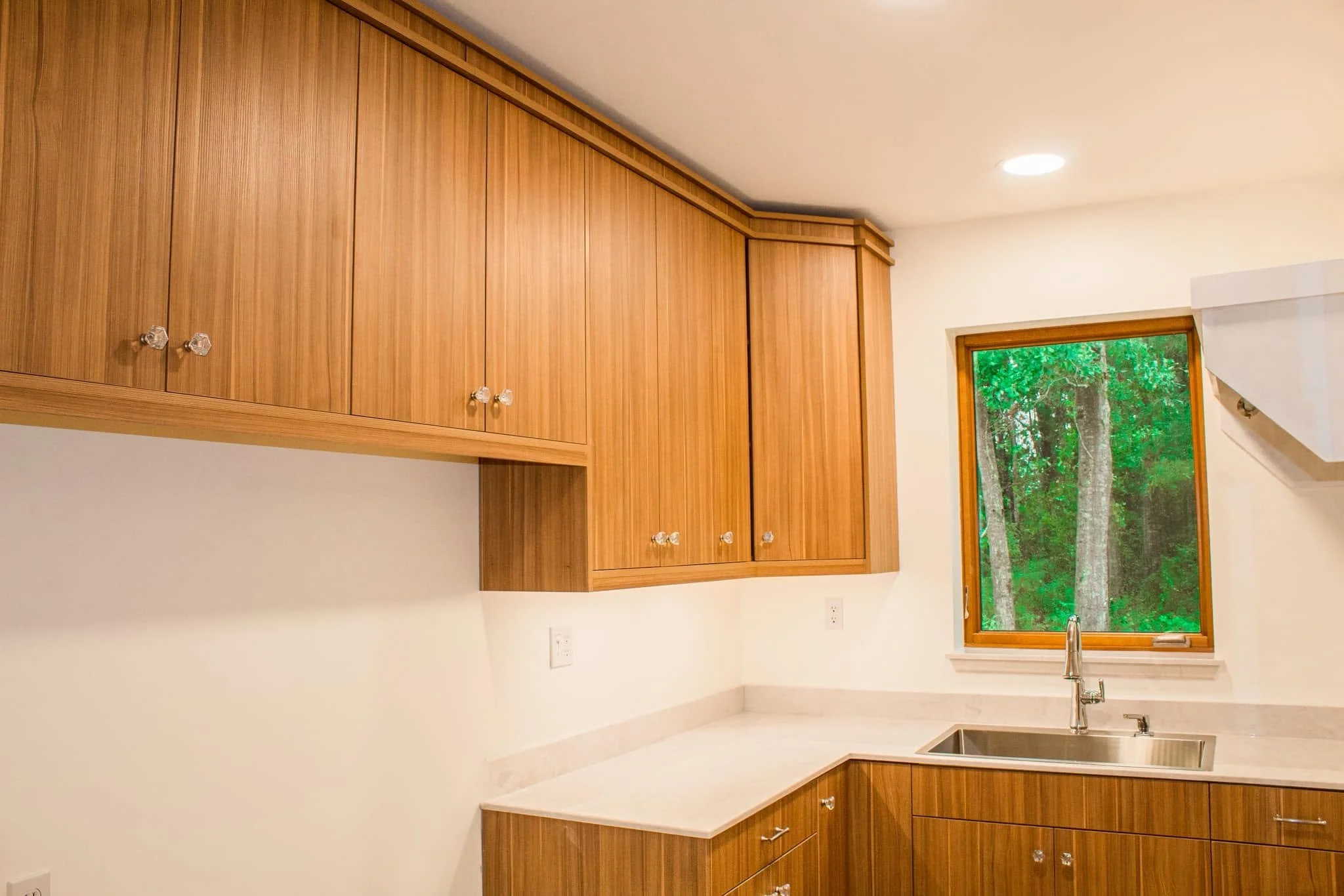 Kitchen with wooden cabinets, white walls, a window showing green trees outside, and a stainless steel sink with a modern faucet.