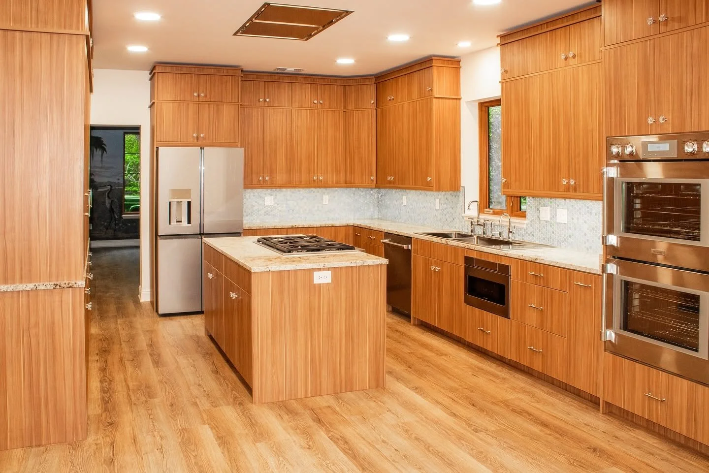 A spacious kitchen with wooden cabinets, granite countertops, a central island with a gas stove, stainless steel appliances including a refrigerator and double oven, a double sink under a window, and hardwood floors.