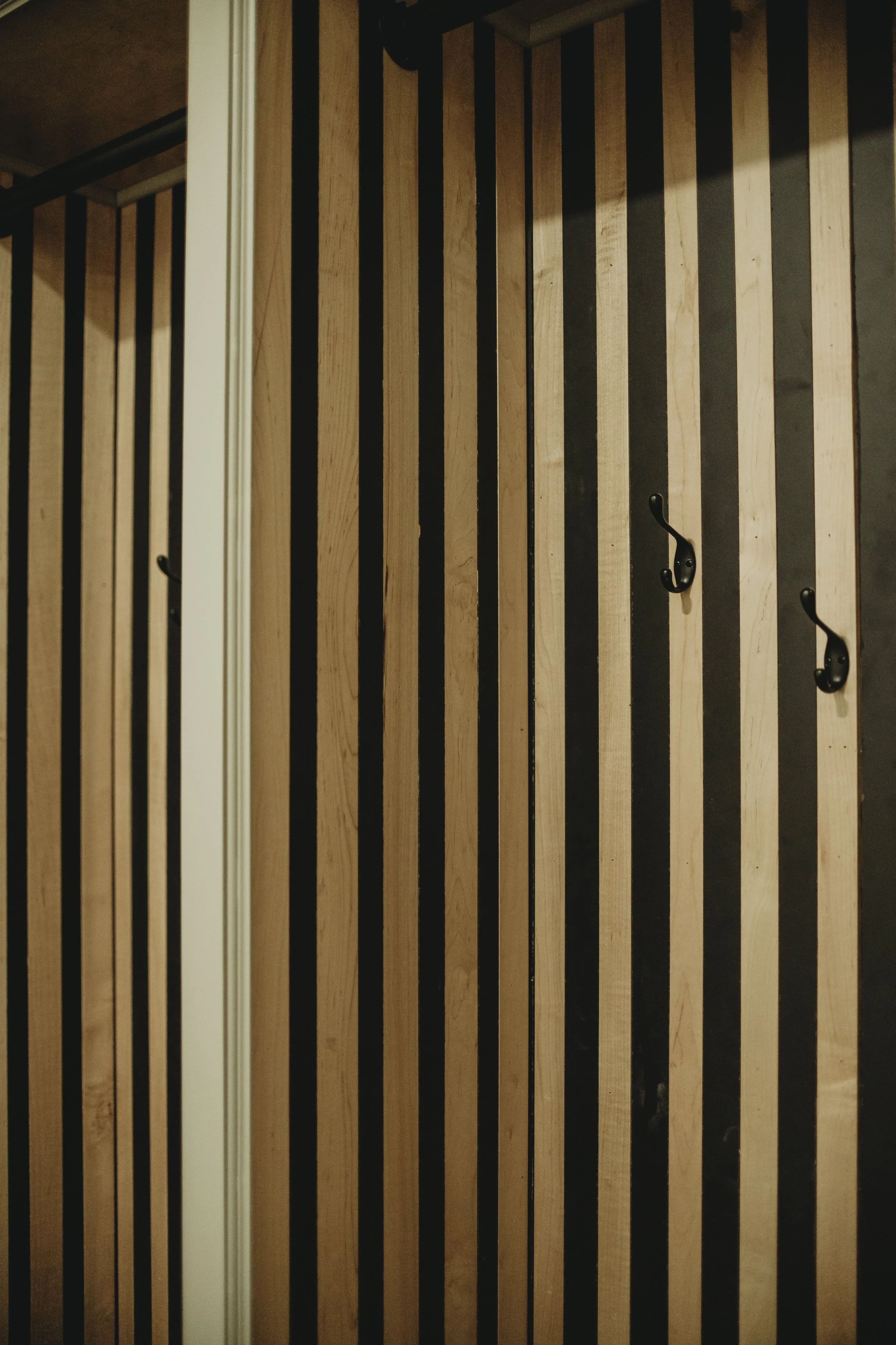 Wooden slat wall with black hooks attached in custom mudroom closet or storage area.