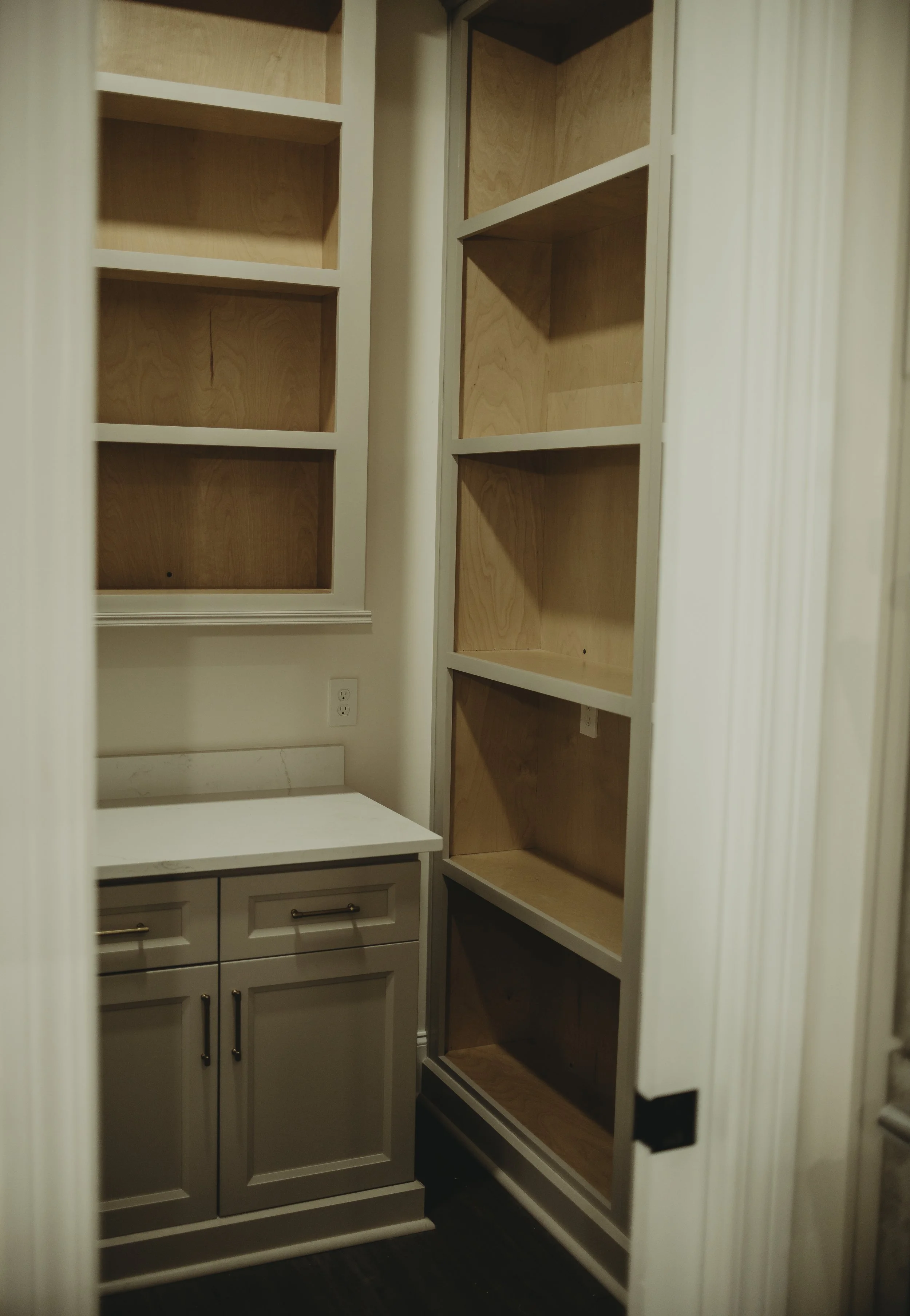 Empty built-in shelves in a small room with a white cabinet and drawers, and a white wall with an electrical outlet.