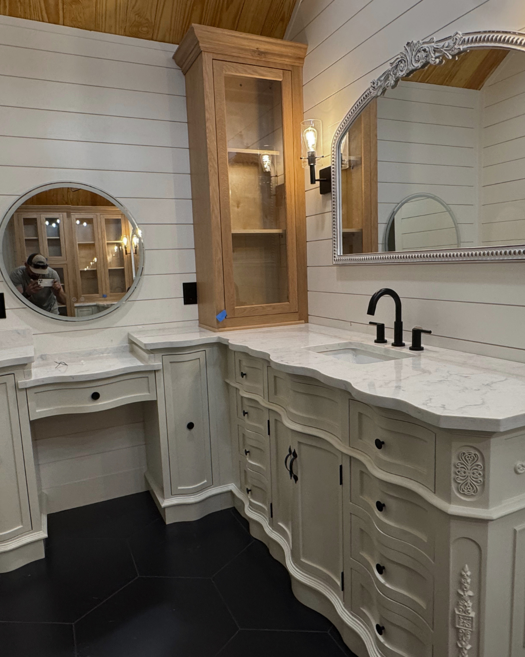 Bathroom vanity with a marble countertop, black faucet, and a mirrored wall. Wooden cabinet with glass door above the counter and shiplap walls.