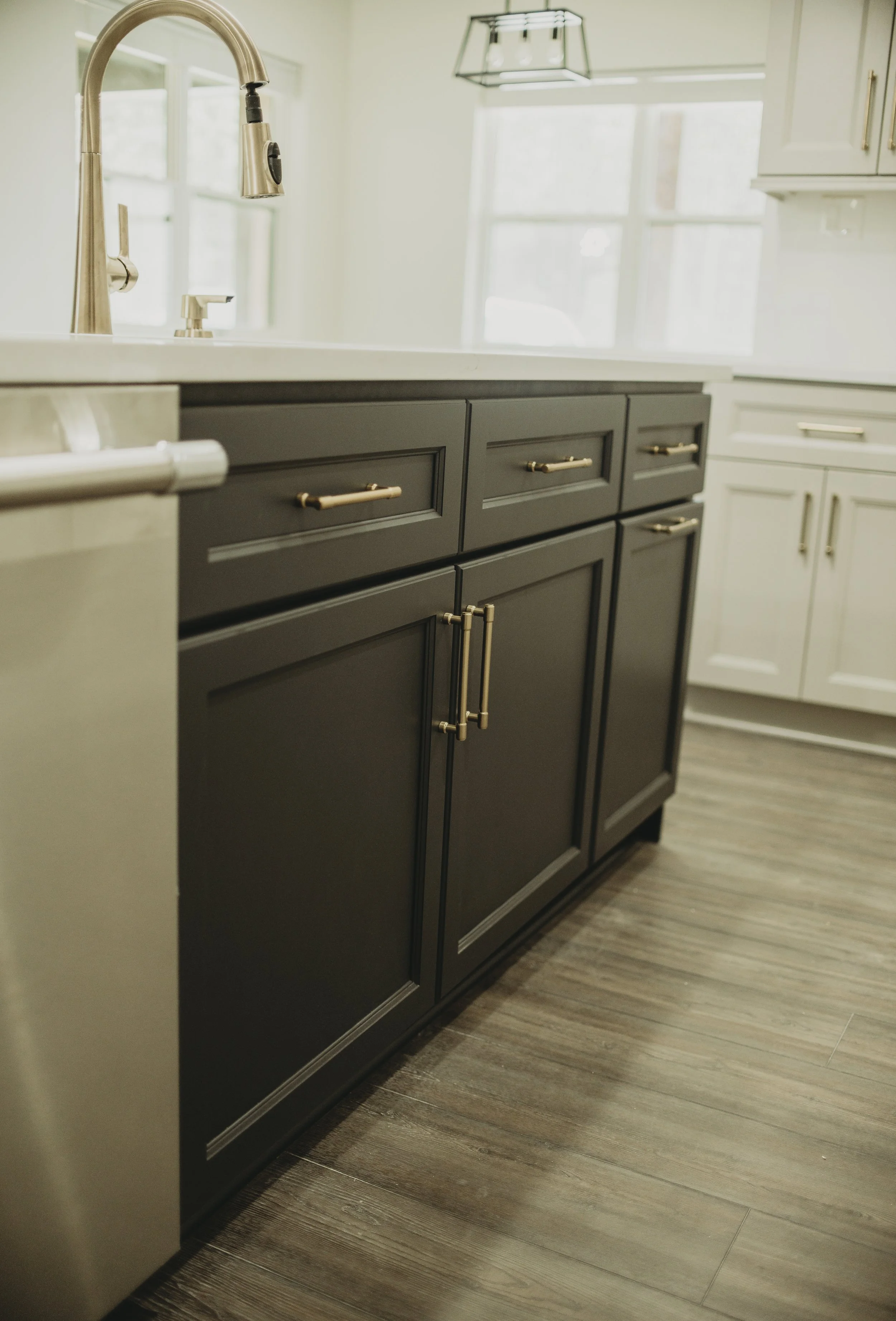 Close-up of a black kitchen cabinet with gold handles, a white countertop, and a brushed stainless steel kitchen faucet. In the background, there are white cabinets and windows with natural light.