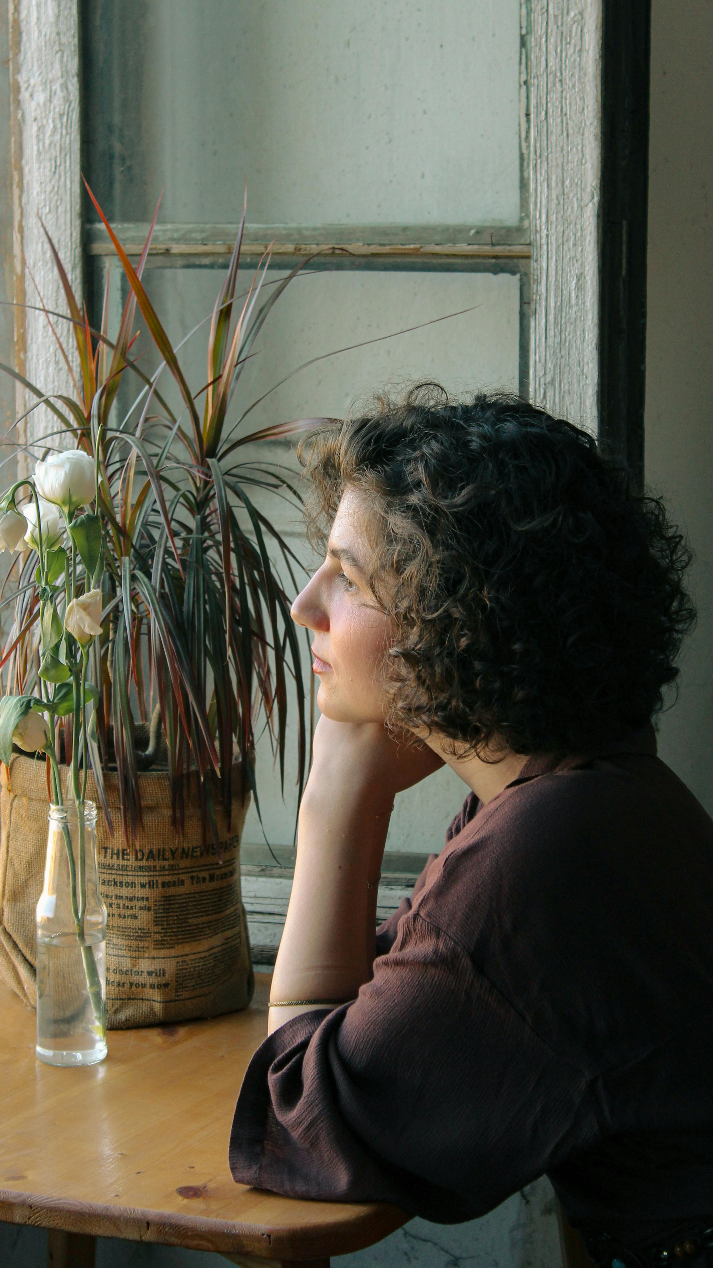 midlife woman with curly hair sits by a window, chin in hand, lost in thought about self abandonment and finding her way back to herself
