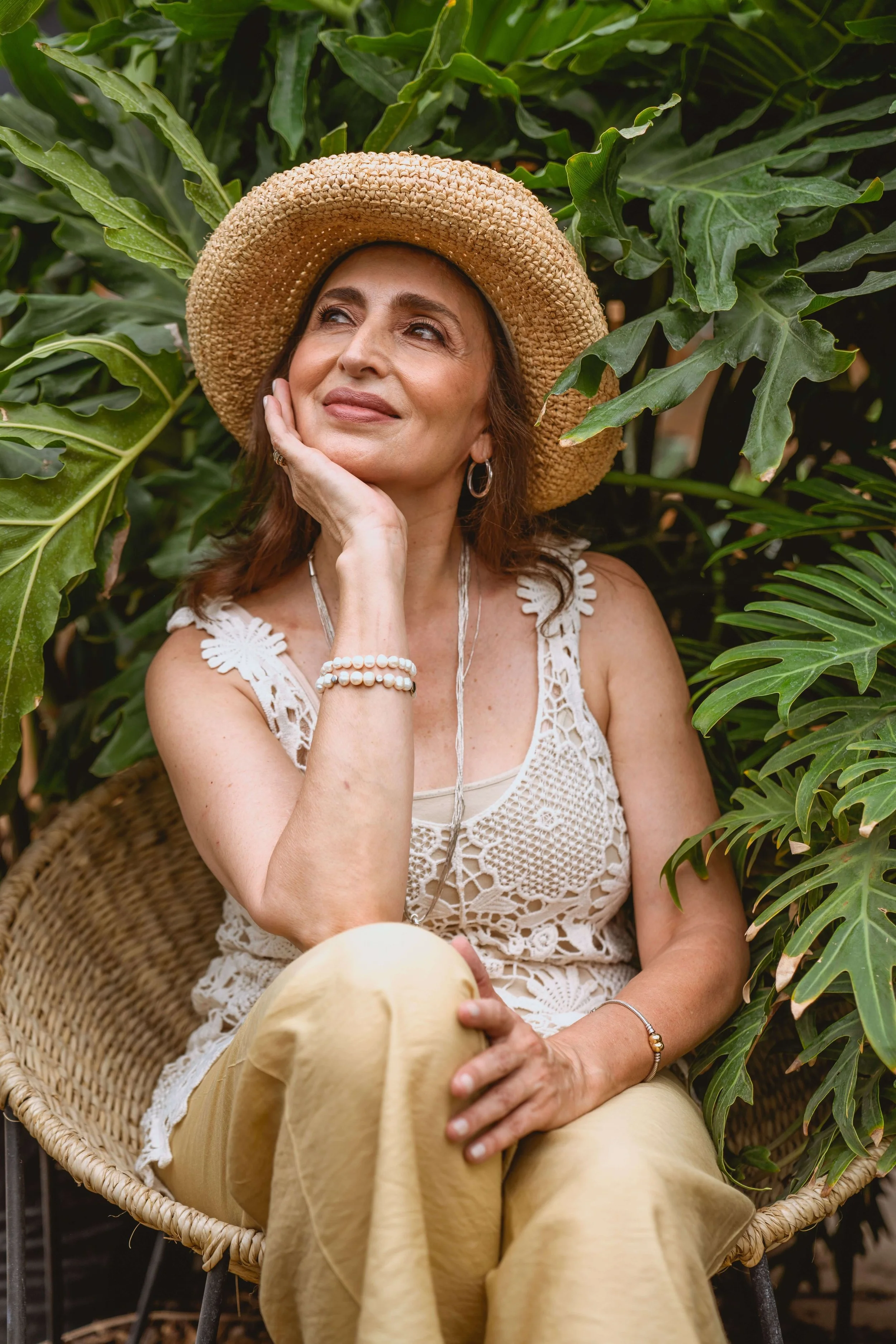 midlife woman in a straw hat smiles softly in a woven chair surrounded by green plants, reflecting the calm of emdr intensive healing