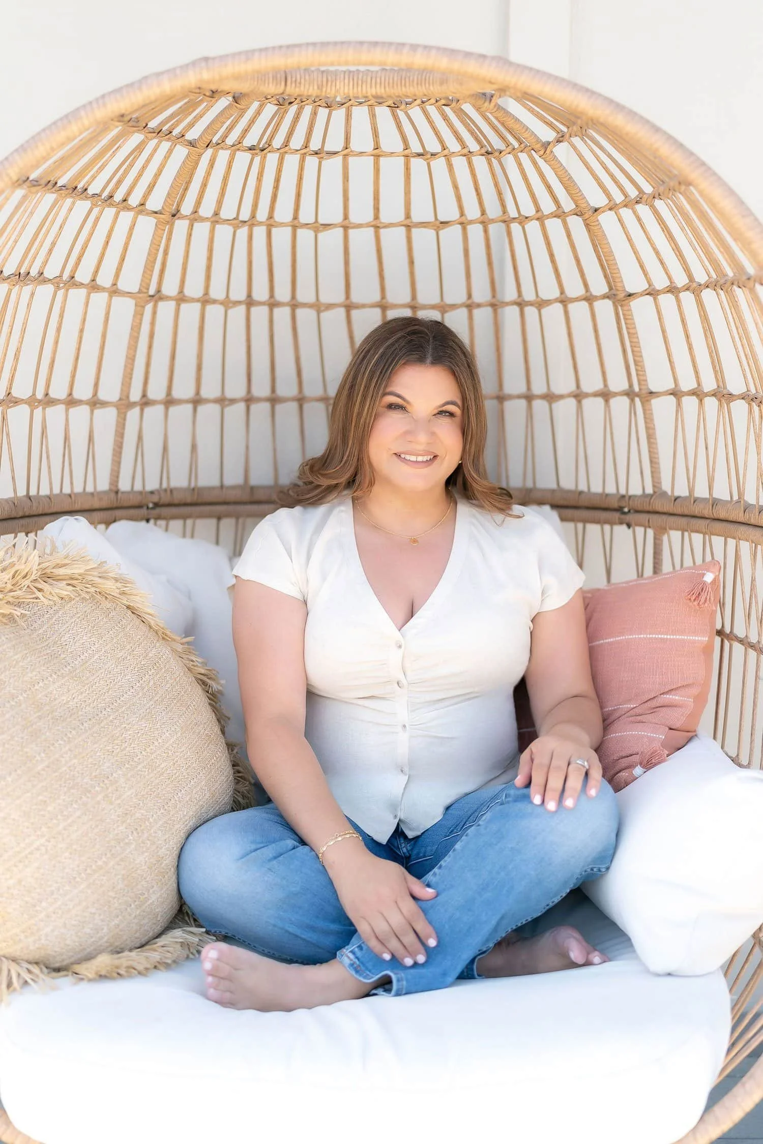 Bicultural woman of color therapist smiling warmly while seated in a woven chair, representing a safe space for healing and navigating guilt.