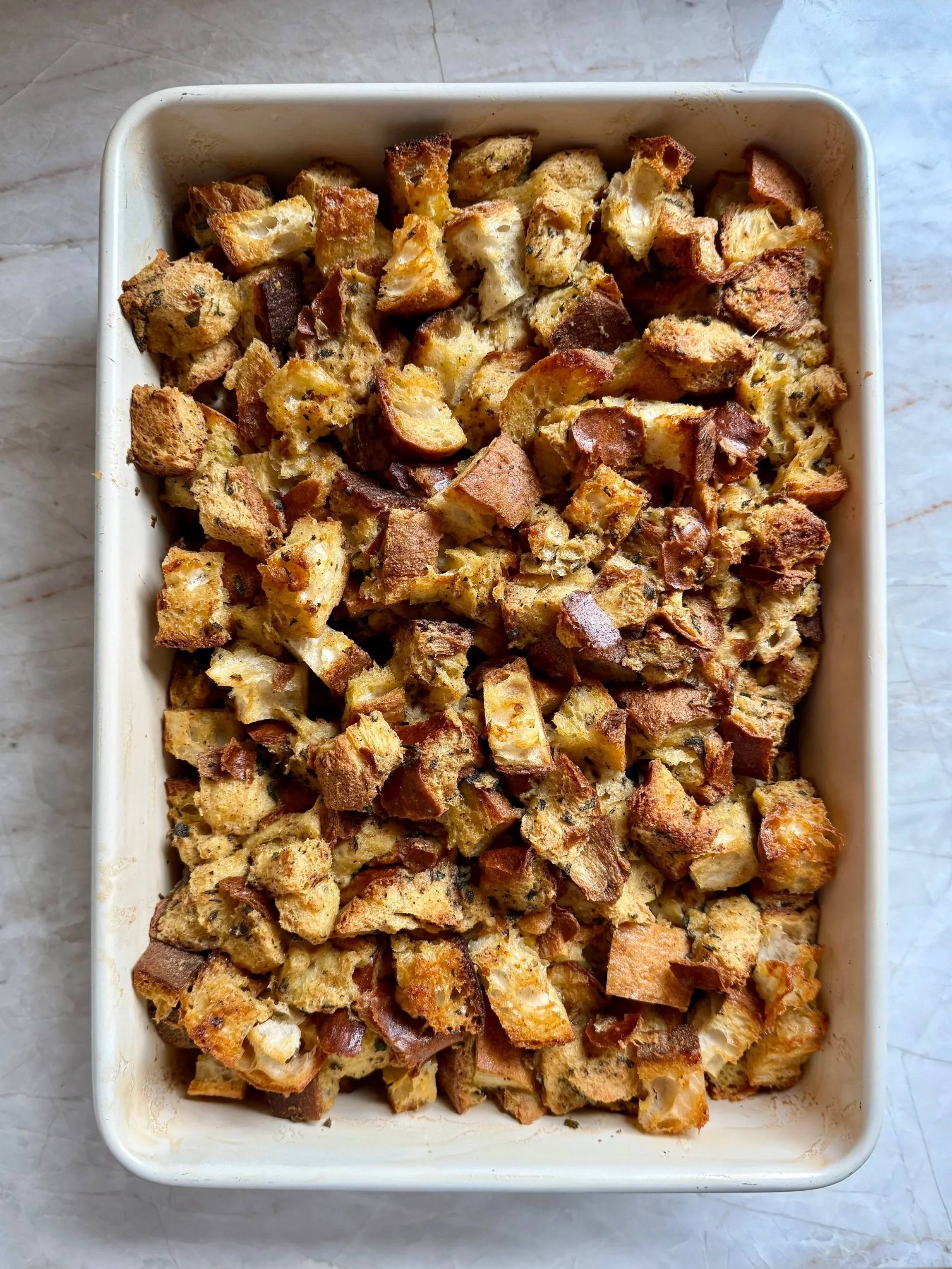 A baking dish filled with golden-brown rustic stuffing made from cubed challah, sourdough and seeded bread, baked until crisp on top with visible herbs throughout.