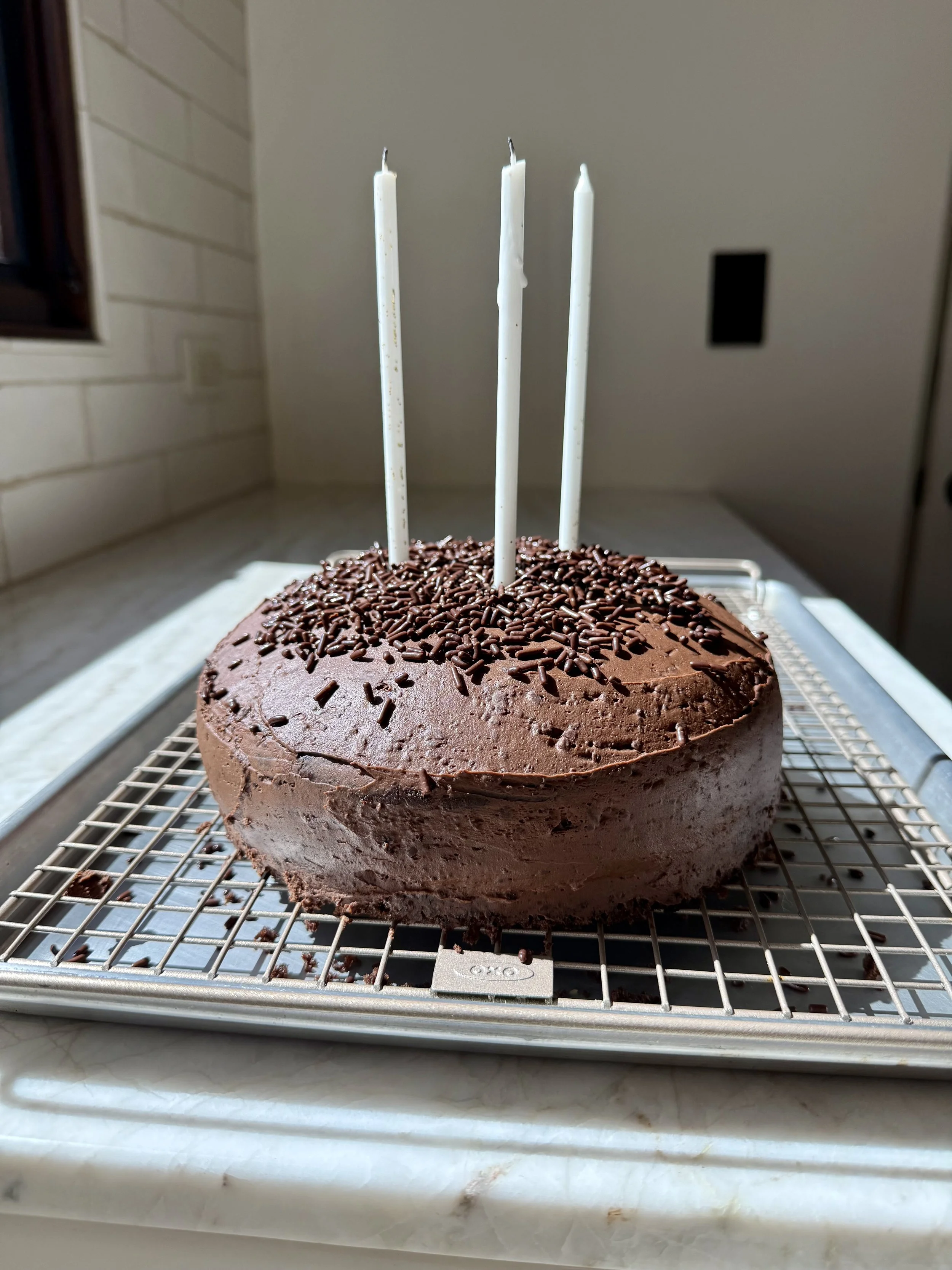 Chocolate peanut butter cup cake topped with chocolate sprinkles and three white candles, sitting on a cooling rack in a sunlit kitchen.