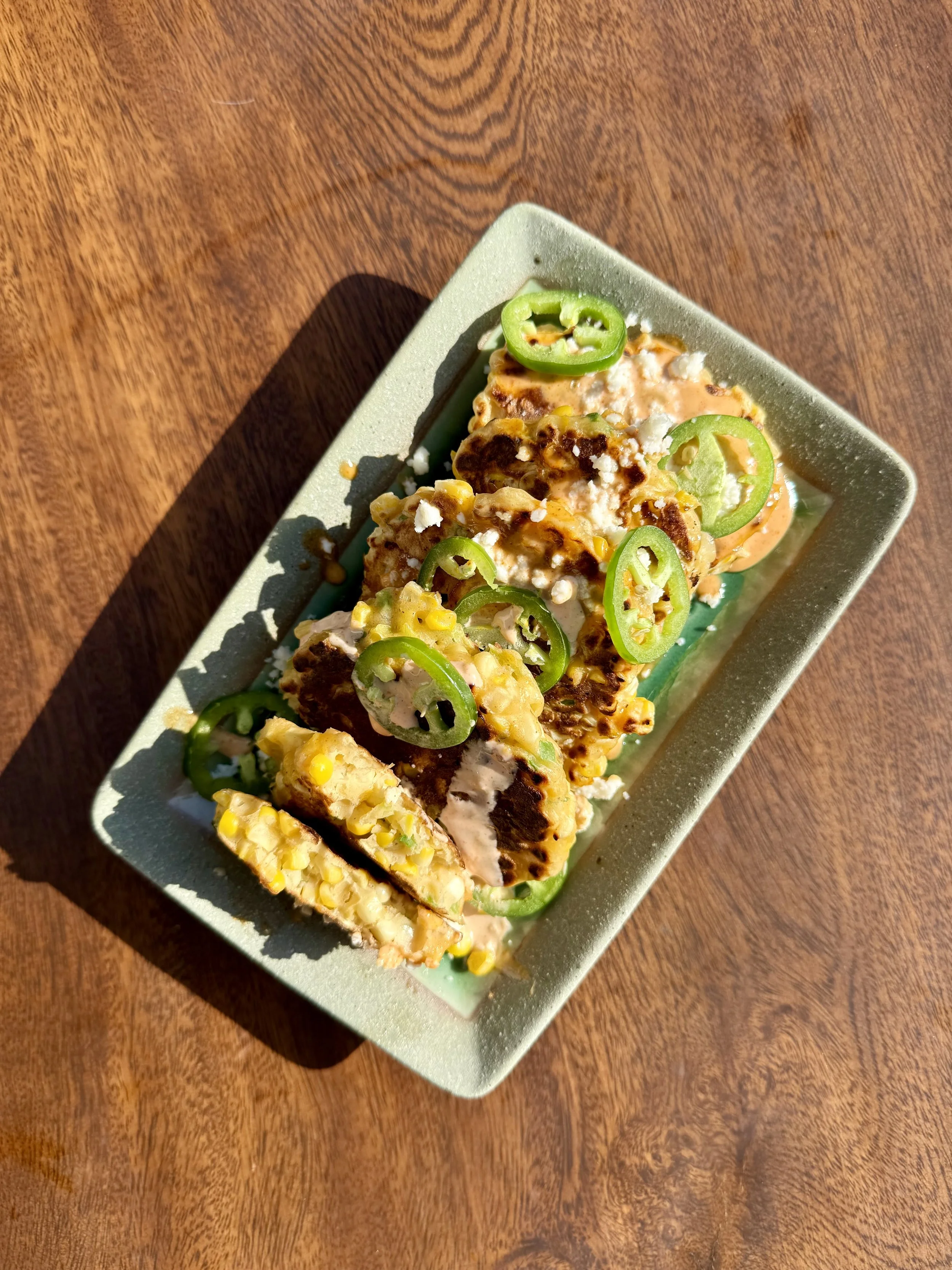 Crispy corn fritters topped with spiced crema, crumbled cotija cheese, and fresh jalapeño slices on a rectangular ceramic serving plate.