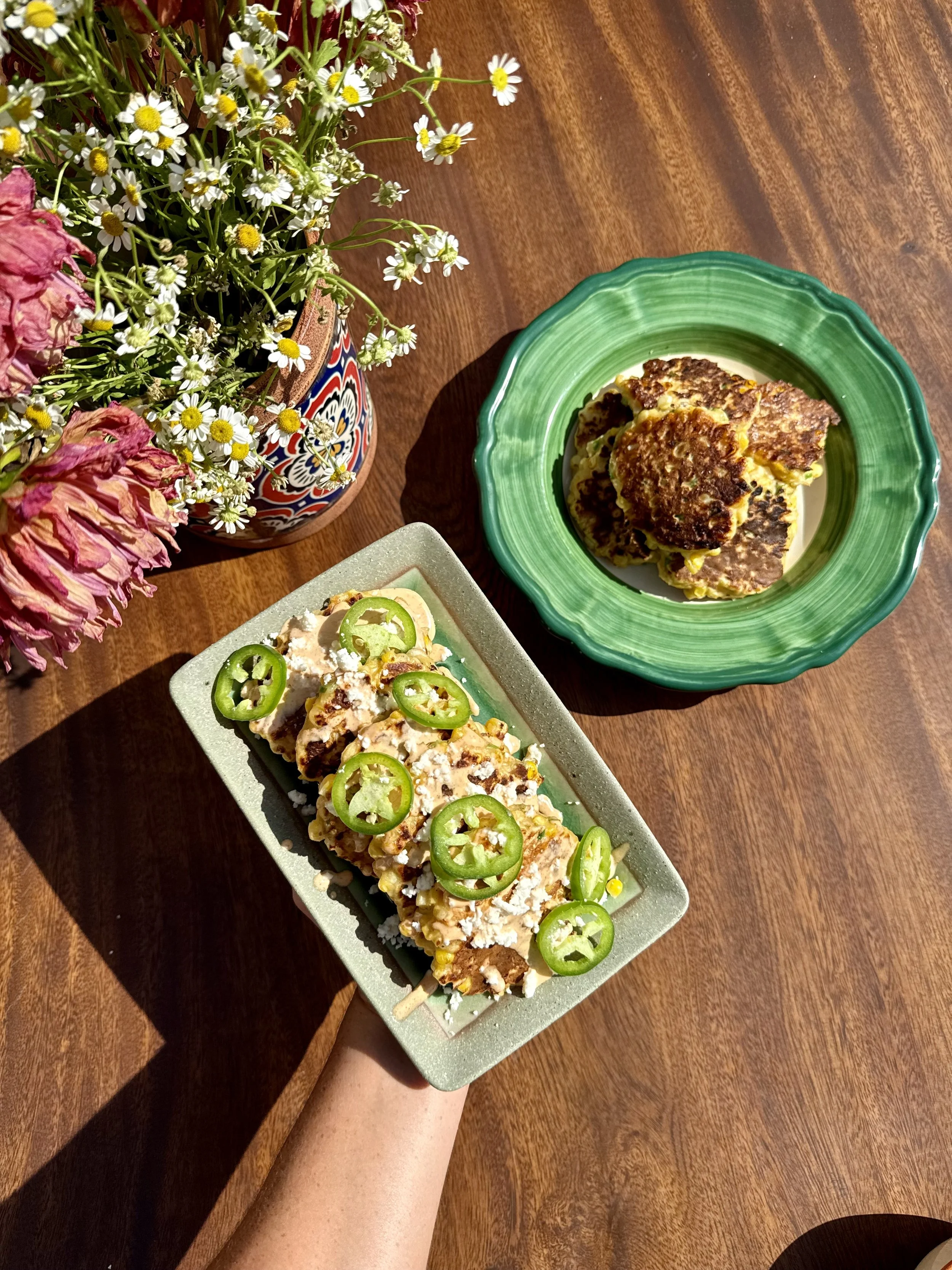 Crispy corn fritters served two ways, with spiced crema, crumbled cotija, and fresh jalapeño slices alongside a plate of golden corn fritters on a sunlit wooden table.
