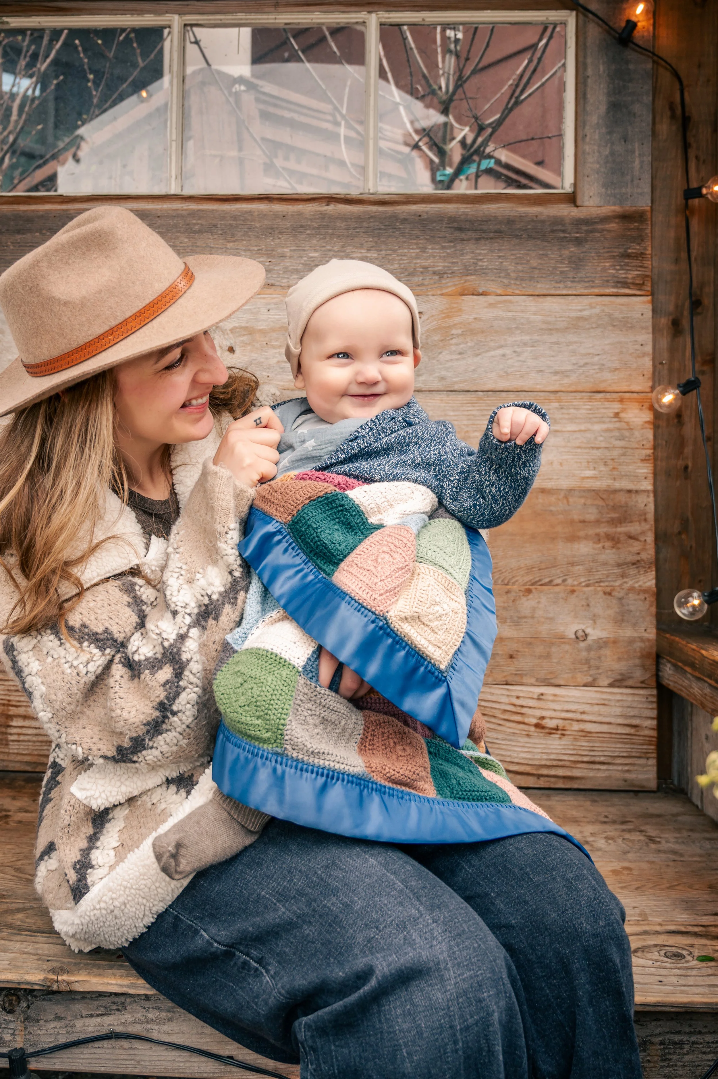 A smiling young woman in a beige hat and cozy sweater sitting on a wooden bench, holding a cheerful baby wrapped in a colorful knitted blanket with a patchwork pattern, against a rustic wooden background with string lights.